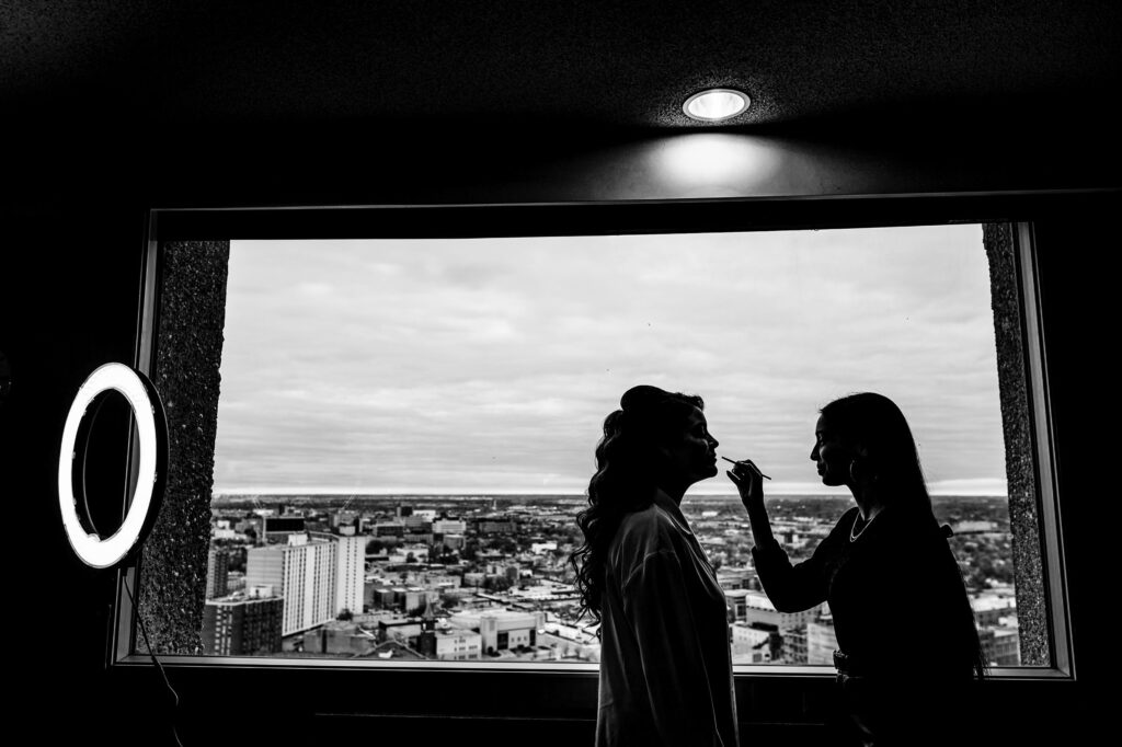 Silhouette of makeup artist preparing for a fall wedding near a large window overlooking the city.