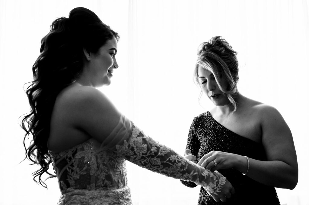 In a dim room, a woman helps a bride with her dress at a fall wedding in Winnipeg.