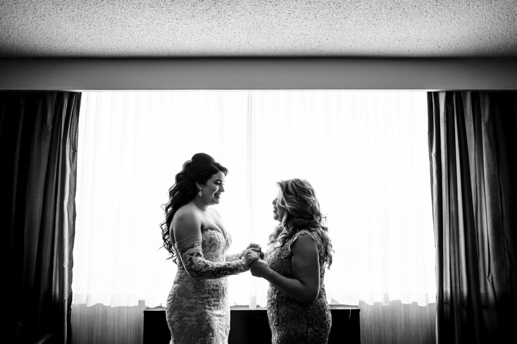 Two women in elegant dresses hold hands, framed by a sunlit window at a fall wedding.