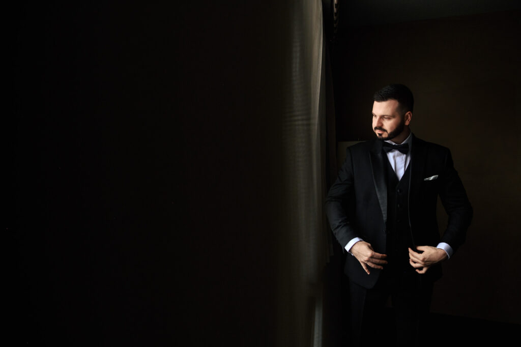 Man in a suit adjusts his jacket by the window at a fall wedding in Winnipeg.
