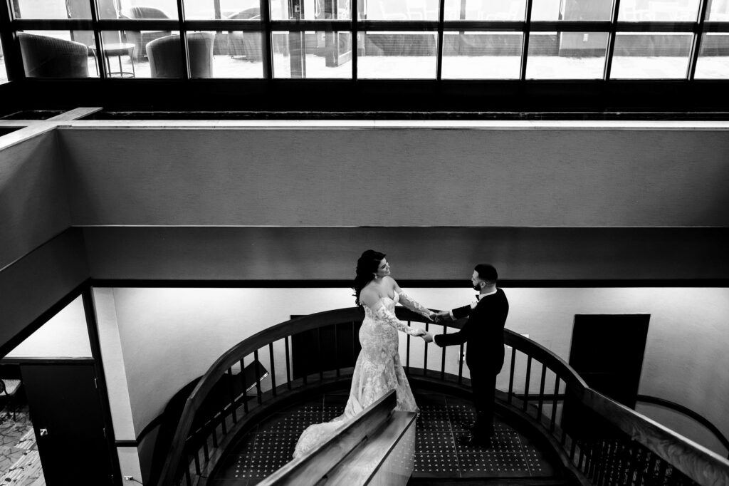 Bride and groom hold hands on a curved staircase at a fall wedding in Winnipeg, framed by large windows.