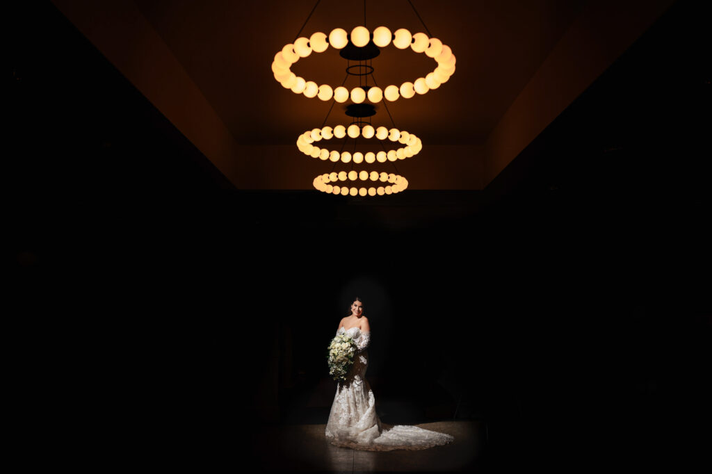 Bride in white dress stands under chandeliers, holding bouquet at fall wedding in Winnipeg.