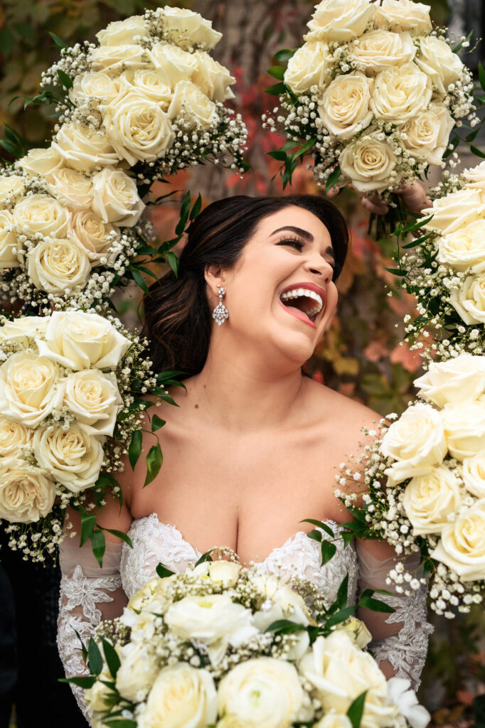 Bride smiling during fall wedding, surrounded by white roses in a lace dress outdoors.