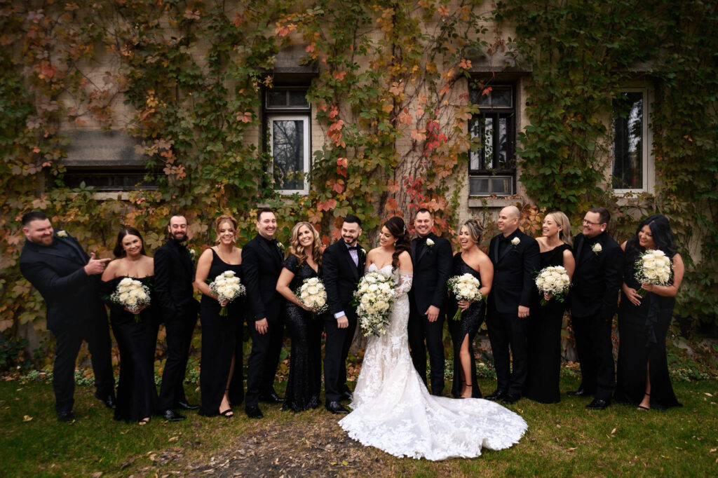 Fall wedding in Winnipeg: Bride in white, black-clad party poses by vine-covered wall outdoors.