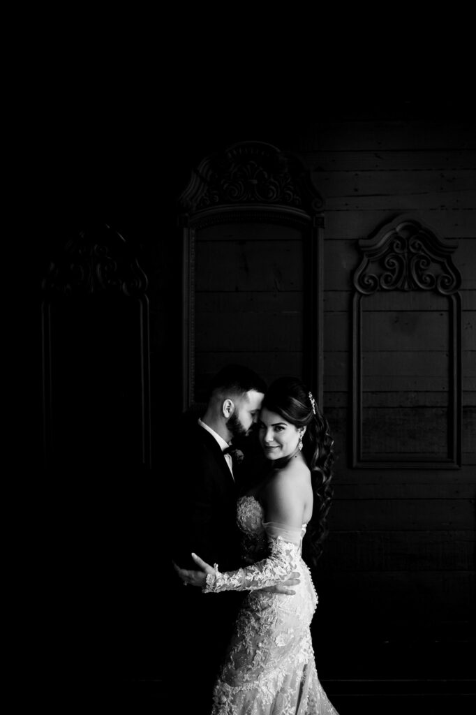 Bride and groom embrace in a dark room; she wears lace, he a tux at their fall wedding.