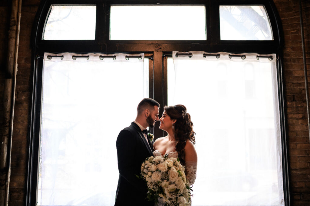 Couple embracing indoors at fall wedding, holding bouquet with light streaming through windows.