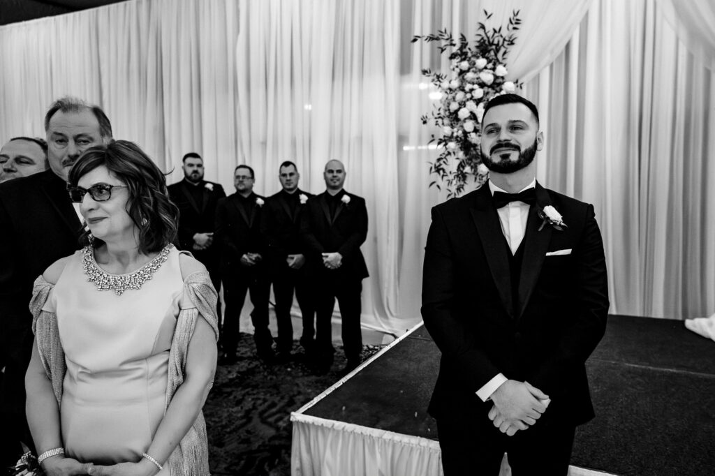 Groom and woman stand together; groomsmen lined up at indoor fall wedding in Winnipeg.
