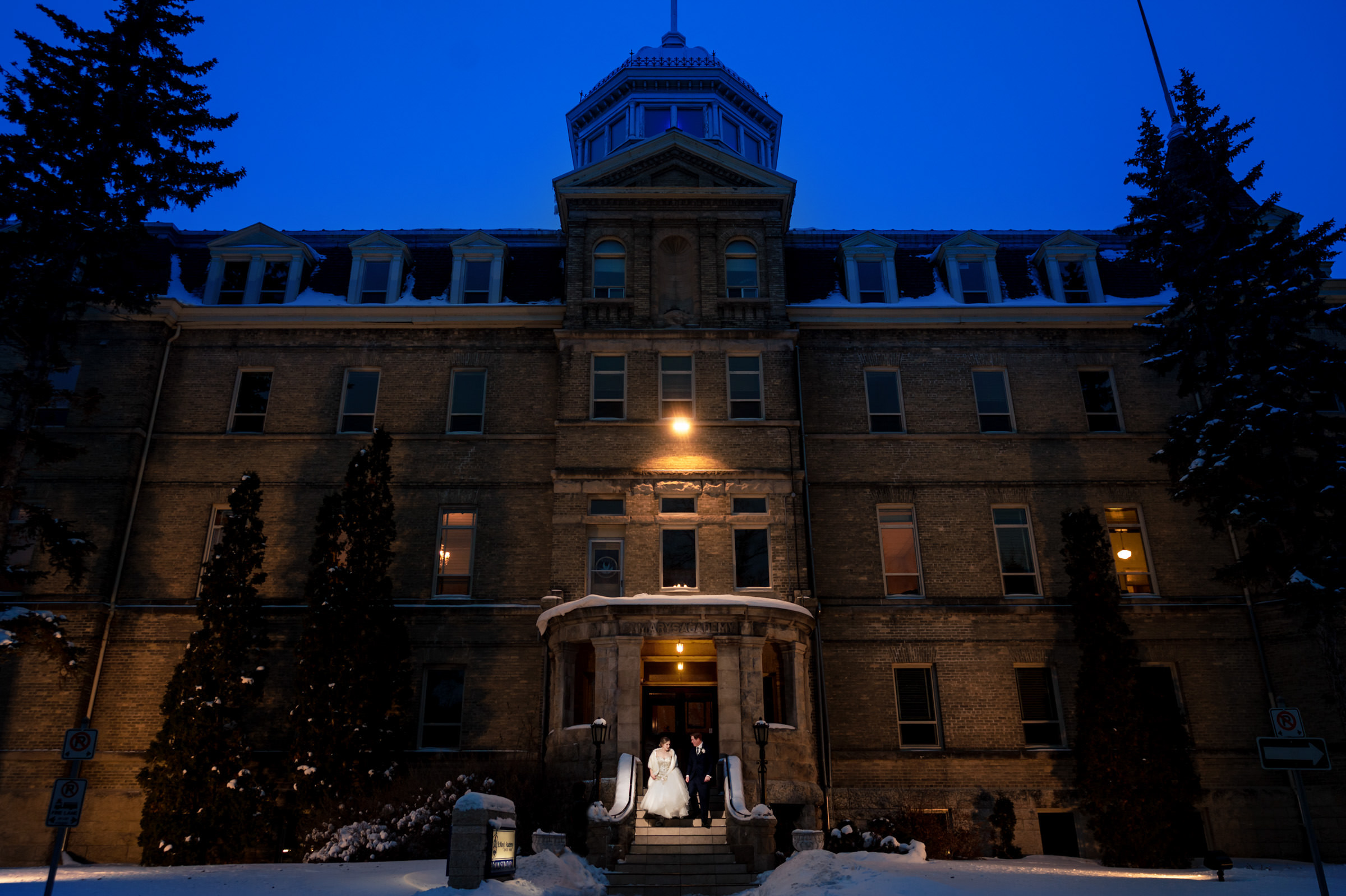 A couple stands on the snowy steps of a large, historic brick building at twilight—the setting for one of their favorite wedding photos. The building is illuminated by soft warm light, highlighting its architectural details and tower. Tall evergreen trees frame the scene under a deep blue sky.