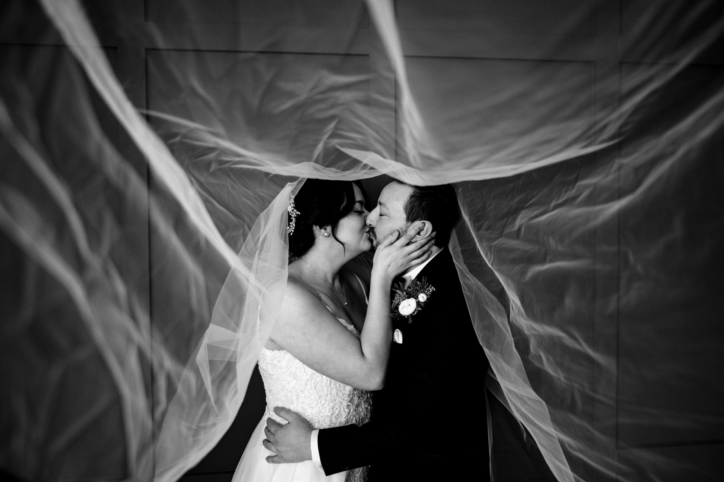 A bride and groom kiss tenderly beneath a flowing veil in one of their favourite wedding photos. The monochrome image captures the romantic moment, with the veil swirling around them, creating a dreamy and intimate atmosphere.