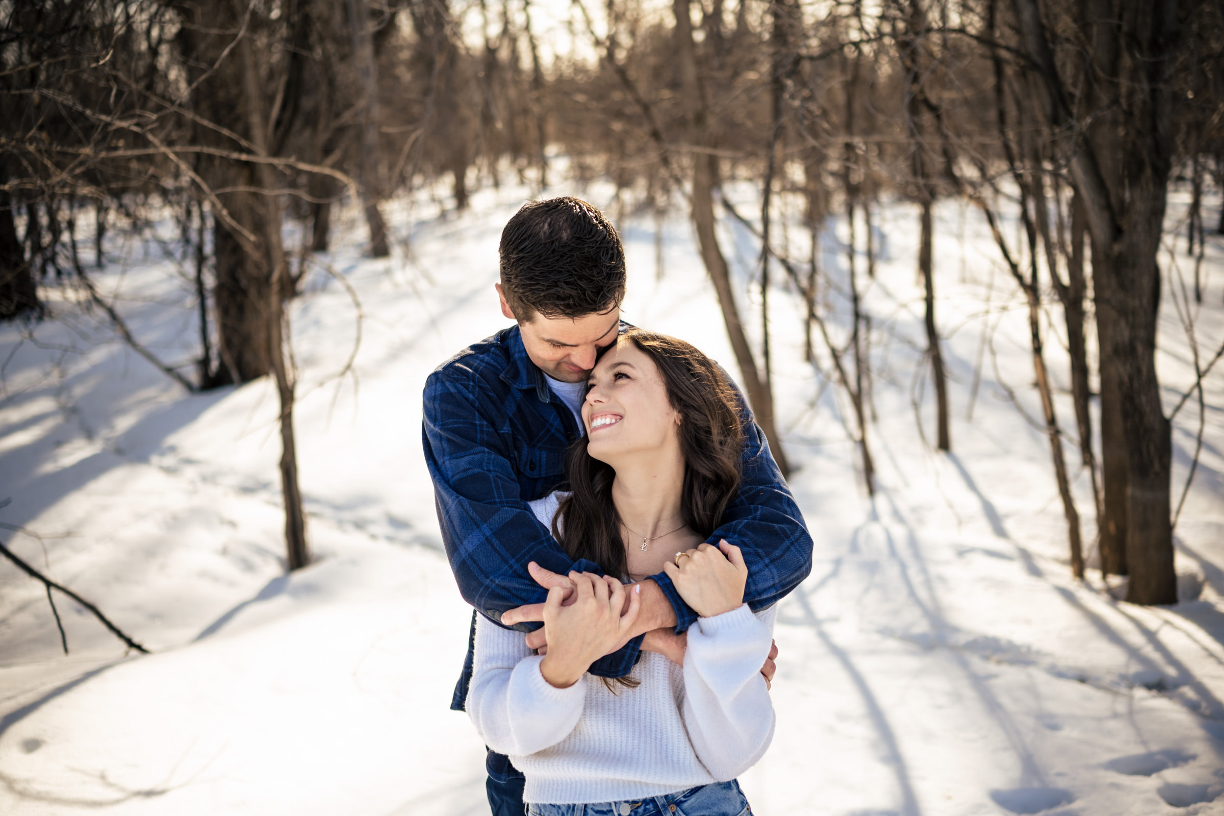 A couple embraces in a snowy forest, creating one of their favorite wedding photos. The man, in a plaid shirt, hugs the woman, in a white sweater, from behind. Sunlight filters through the bare trees, casting shadows on the snow as they smile warmly and affectionately.