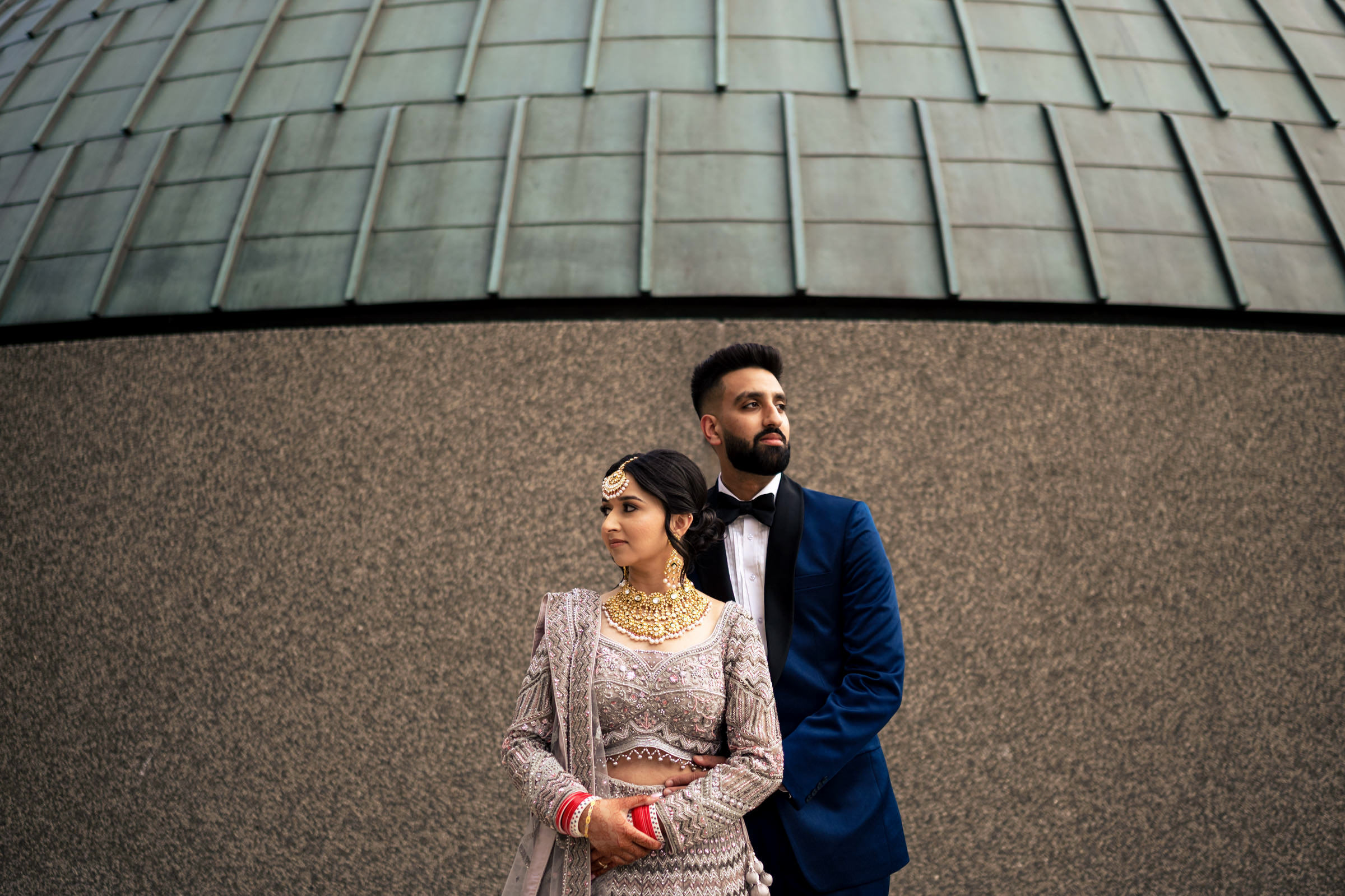 A couple dressed in formal attire stands in front of a modern, textured building. The woman wears an ornate, traditional outfit with jewelry, while the man sports a royal blue suit and black bow tie. Both gaze to the side, creating one of their favourite wedding photos.