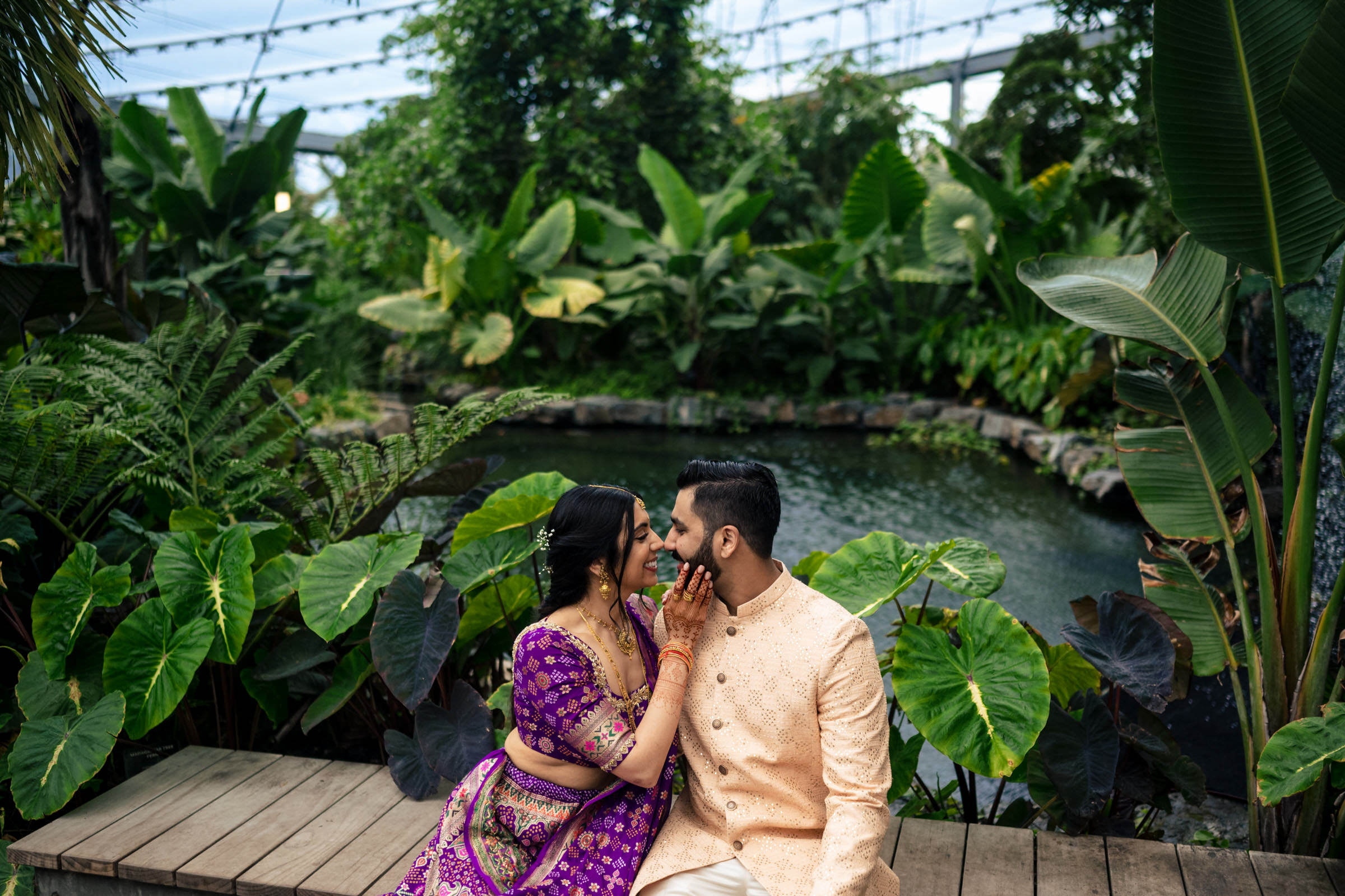 A couple sits on a wooden deck in a lush garden, large green leaves and a pond framing the scene. The woman, in a purple and gold sari, lovingly holds the man's face as they smile at each other in their favourite wedding photos, both dressed in traditional attire.