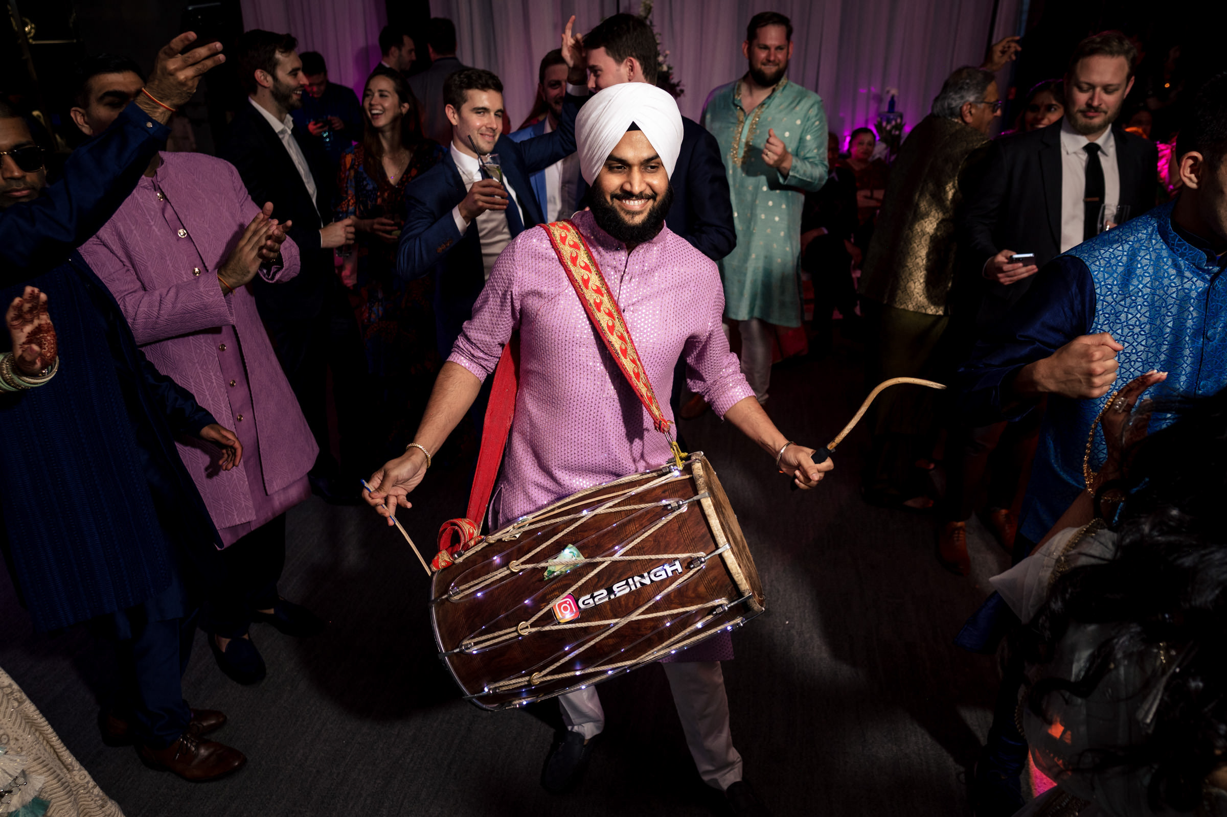A man in a pink kurta and white turban joyfully plays a dhol drum at a lively gathering, captured in one of my favorite wedding photos. People clap and smile around him, creating a festive atmosphere under the dimly lit, colorful lights.