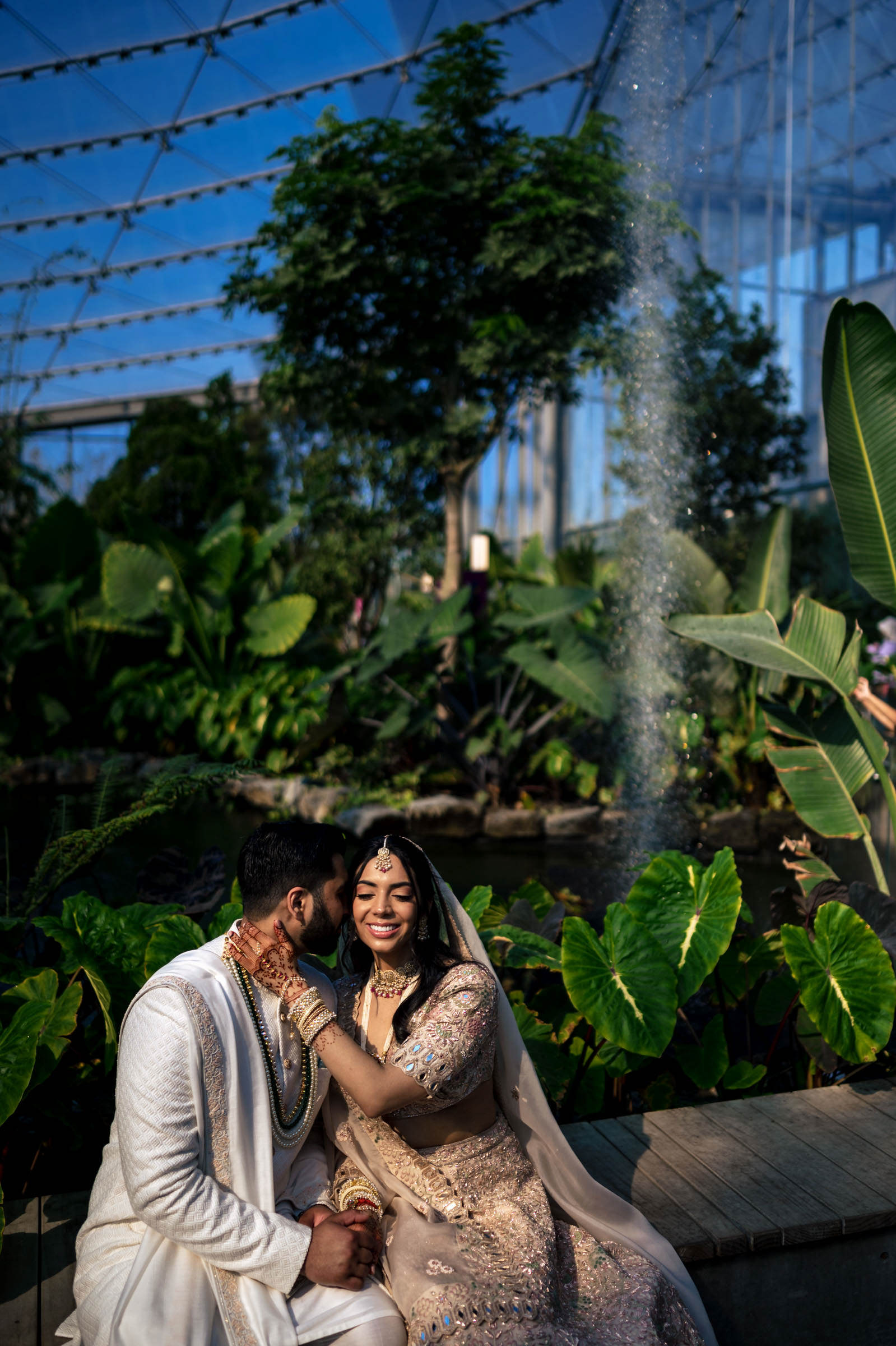 A couple in traditional attire sits joyfully in a lush indoor garden, surrounded by greenery. The woman touches the man's face, both smiling under a sunlit glass canopy with a tree and large leaves in the background, creating one of their favourite wedding photos.