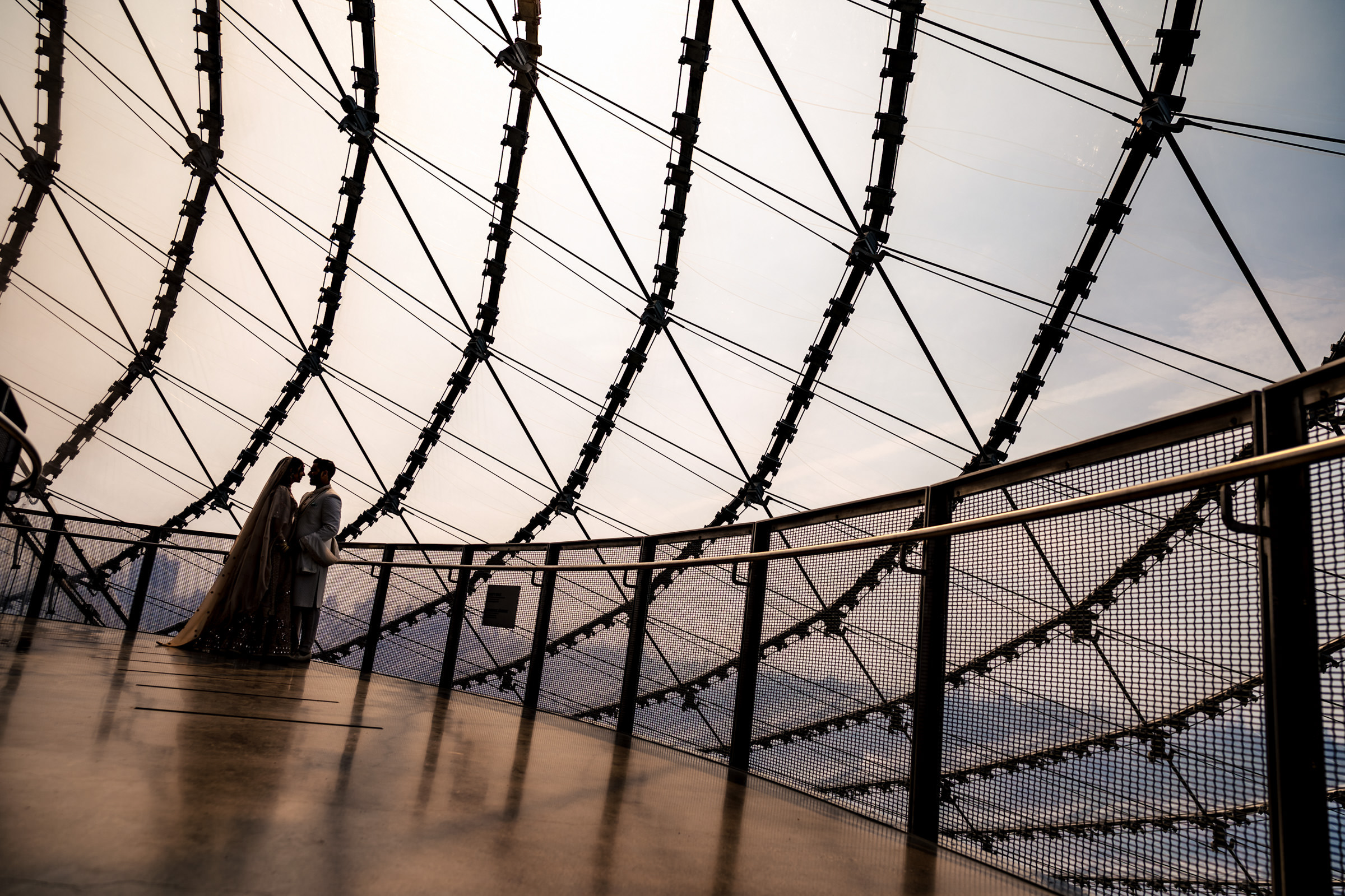 Two people stand together on a modern lookout platform, capturing one of their favourite wedding photos. The metal and wire structures frame their view as they gaze outward, with a dramatic sky in the background, creating a striking and atmospheric scene.