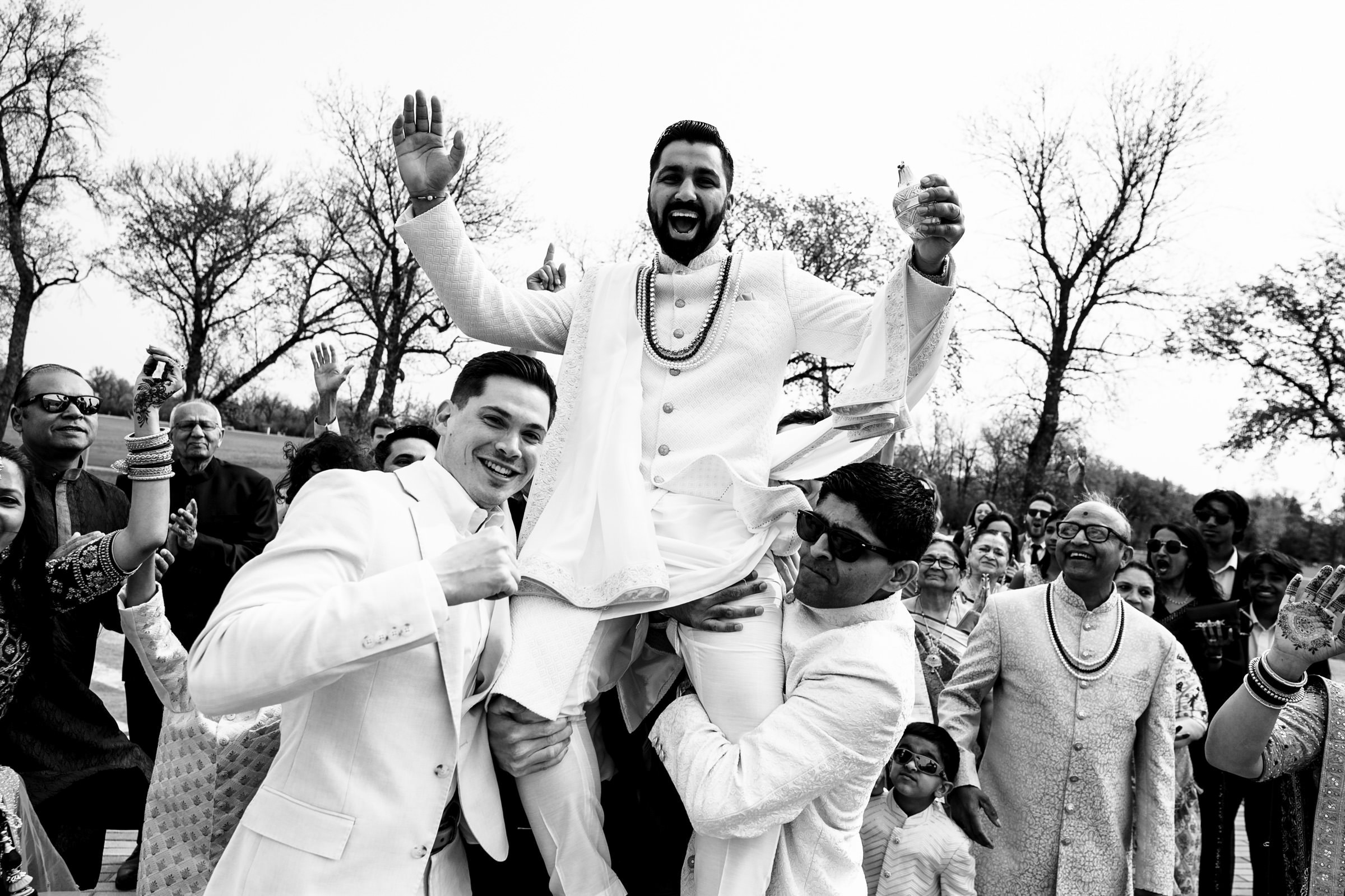 A joyous groom dressed in traditional attire is lifted up by two men during an outdoor celebration, creating one of the favourite wedding photos of the day. A crowd of smiling guests surrounds them, with trees in the background. The scene is festive and lively.