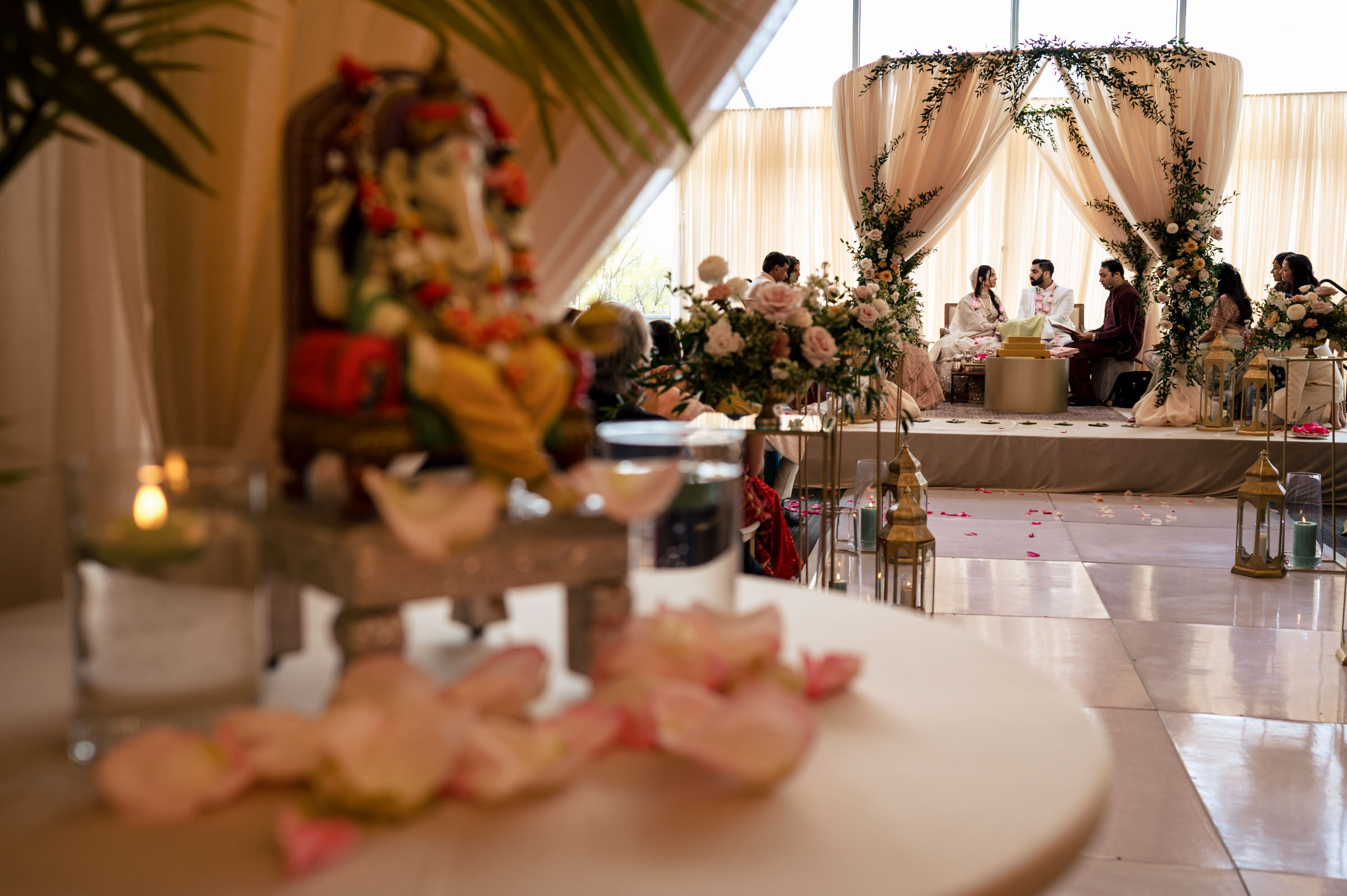 A wedding ceremony unfolds indoors, the couple seated under a floral arch, captured in what will surely become some of their favourite wedding photos. Guests sit on either side, with a small Ganesh statue adorned in rose petals nearby. The elegantly decorated setting features flowers and drapes.