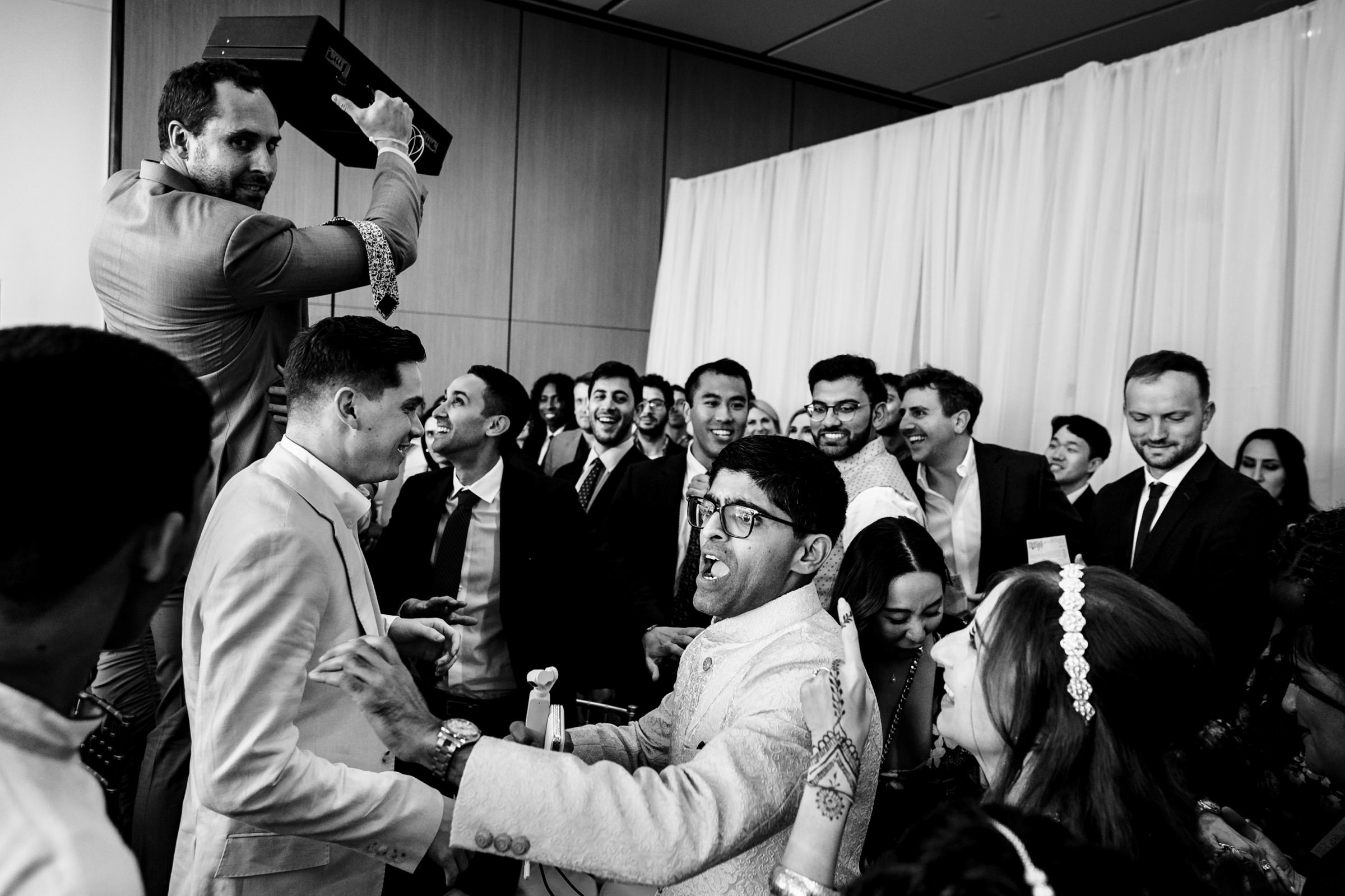 A lively black and white scene at a celebration, reminiscent of favourite wedding photos, with people smiling and interacting. One person holds a box above their head as others gather closely, capturing a joyful and energetic moment against a backdrop of curtains and wooden panels.