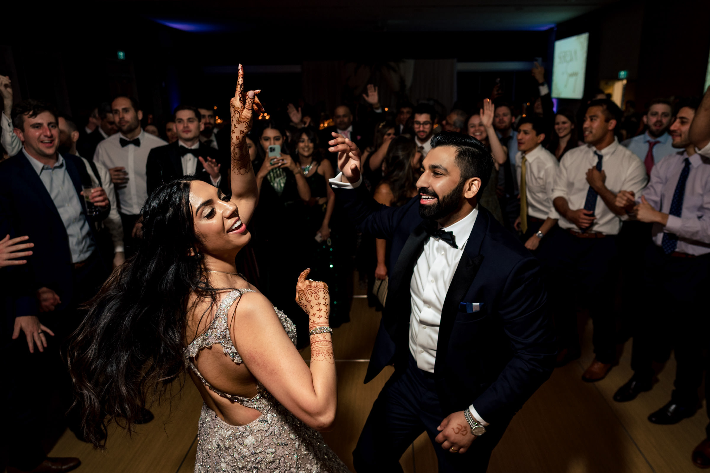 A couple is joyfully dancing at a celebration, the woman in a sleeveless dress and the man in a suit with a bowtie. Surrounded by partygoers clapping along, this moment captures the delight reminiscent of everyone's favourite wedding photos.