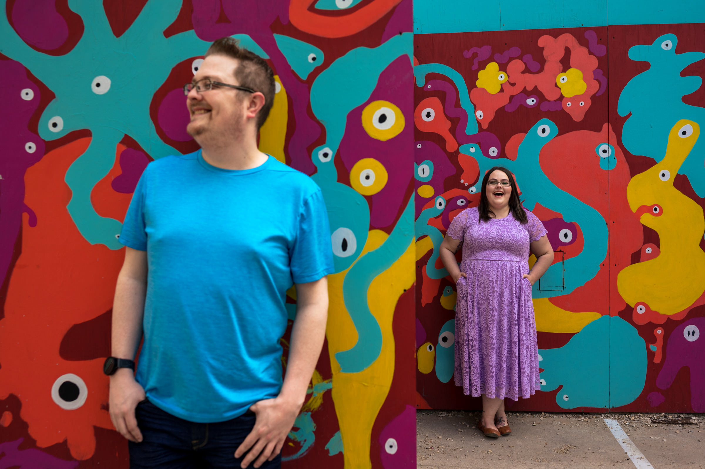 A man in a blue shirt and a woman in a purple dress pose for their favorite wedding photos, standing before a colorful, abstract mural with eyes and vibrant shapes. The man smiles, looking left, while the woman laughs from further back.