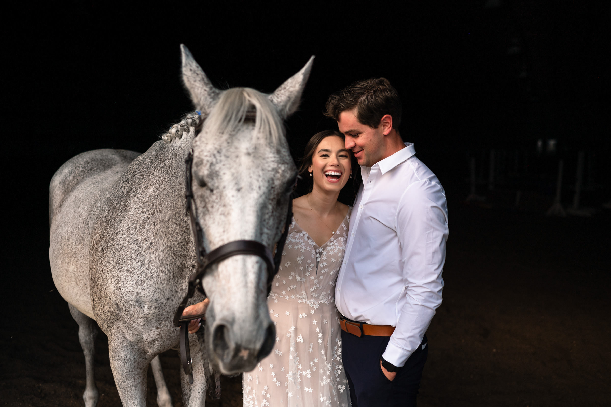 A smiling couple stands close together with a dapple gray horse beside them in one of their favorite wedding photos. The woman wears a delicate, sheer dress with floral patterns, while the man is in a crisp white shirt. The dark background highlights the trio beautifully.