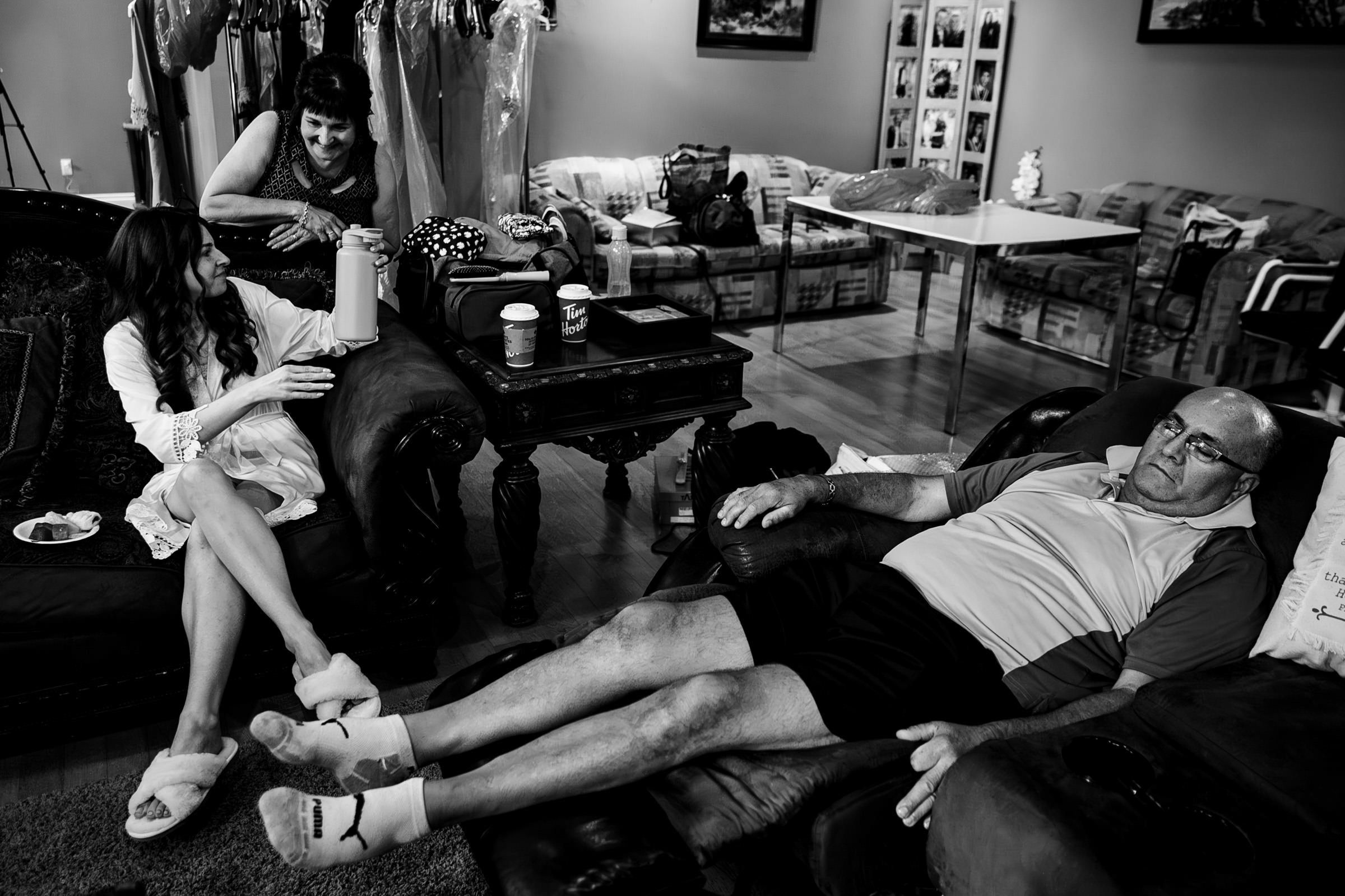 A black and white image of a cozy living room resembles a scene from someone's favourite wedding photos. A woman with long hair sits on a sofa, handing a drink to another standing nearby. A man lies relaxed on a sofa, as clothes hang in the background, and a coffee table completes the scene.