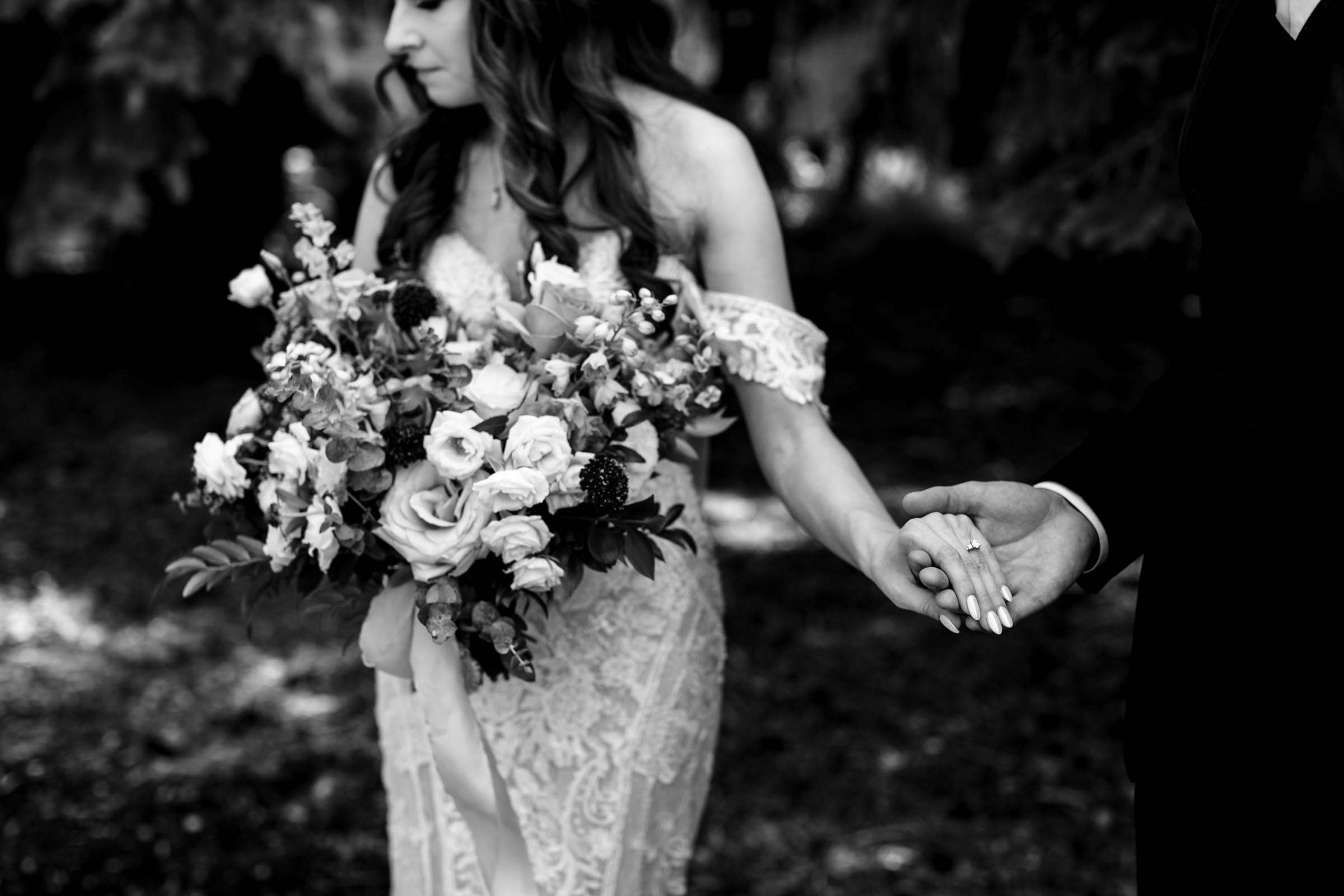 A black and white photo captures one of my favorite wedding moments: a bride in a lace dress, bouquet in hand, looks down as the groom extends his hand towards hers. They're gently enveloped by lush foliage, crafting an intimate and timeless scene.