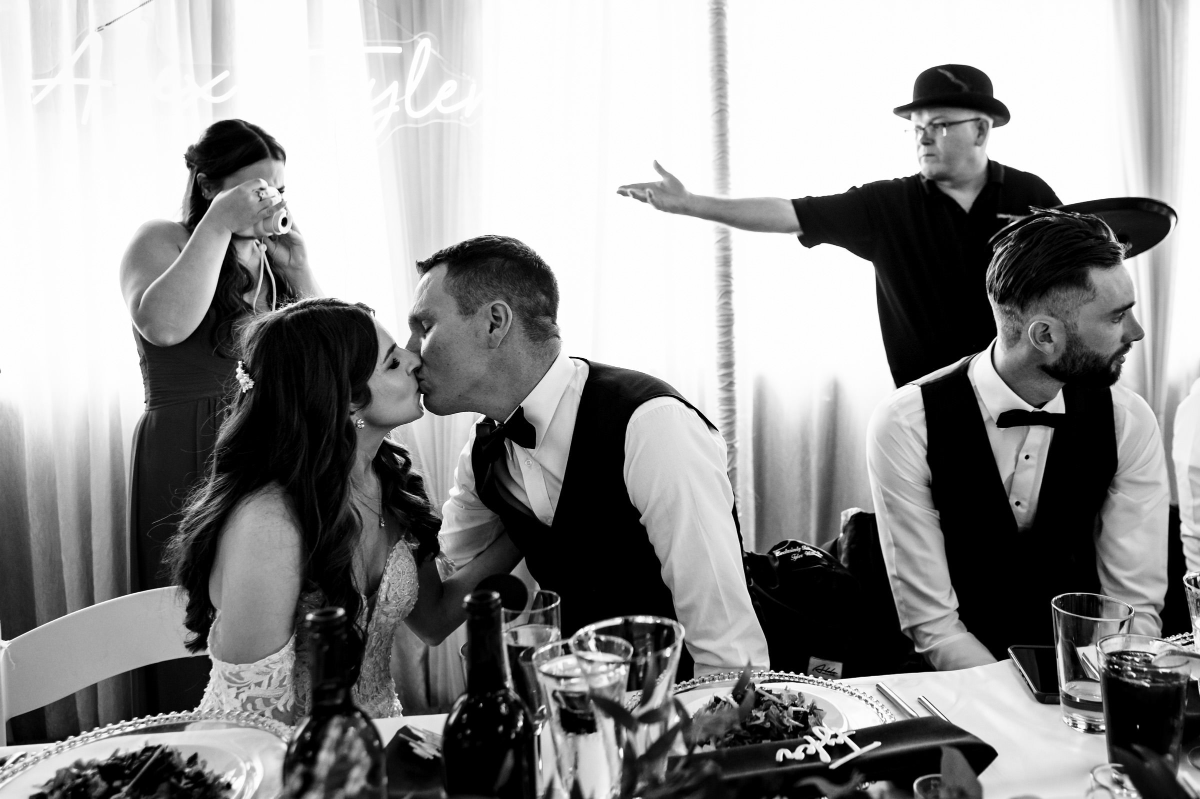 A bride and groom share a kiss at a table, captured among their favorite wedding photos. A man in the background gestures with his arm while a woman snaps the moment on her phone. Another man in formal attire sits to the right, looking away from the festive scene.