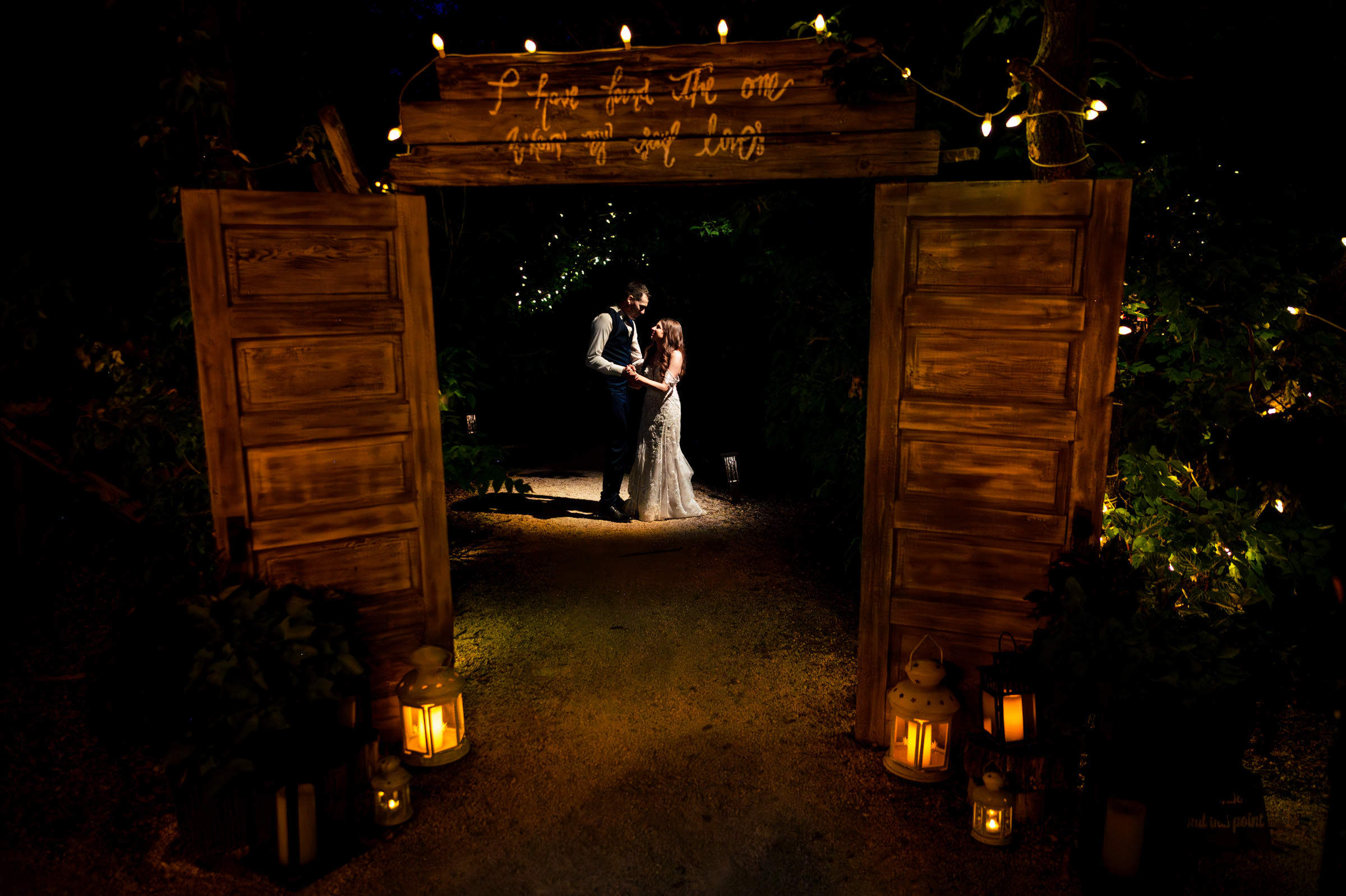 A couple dances under a wooden archway adorned with string lights in a dimly lit, rustic setting. Lanterns with candles illuminate the path, capturing one of their favourite wedding photos. The warm glow creates a romantic atmosphere against the dark background of trees.