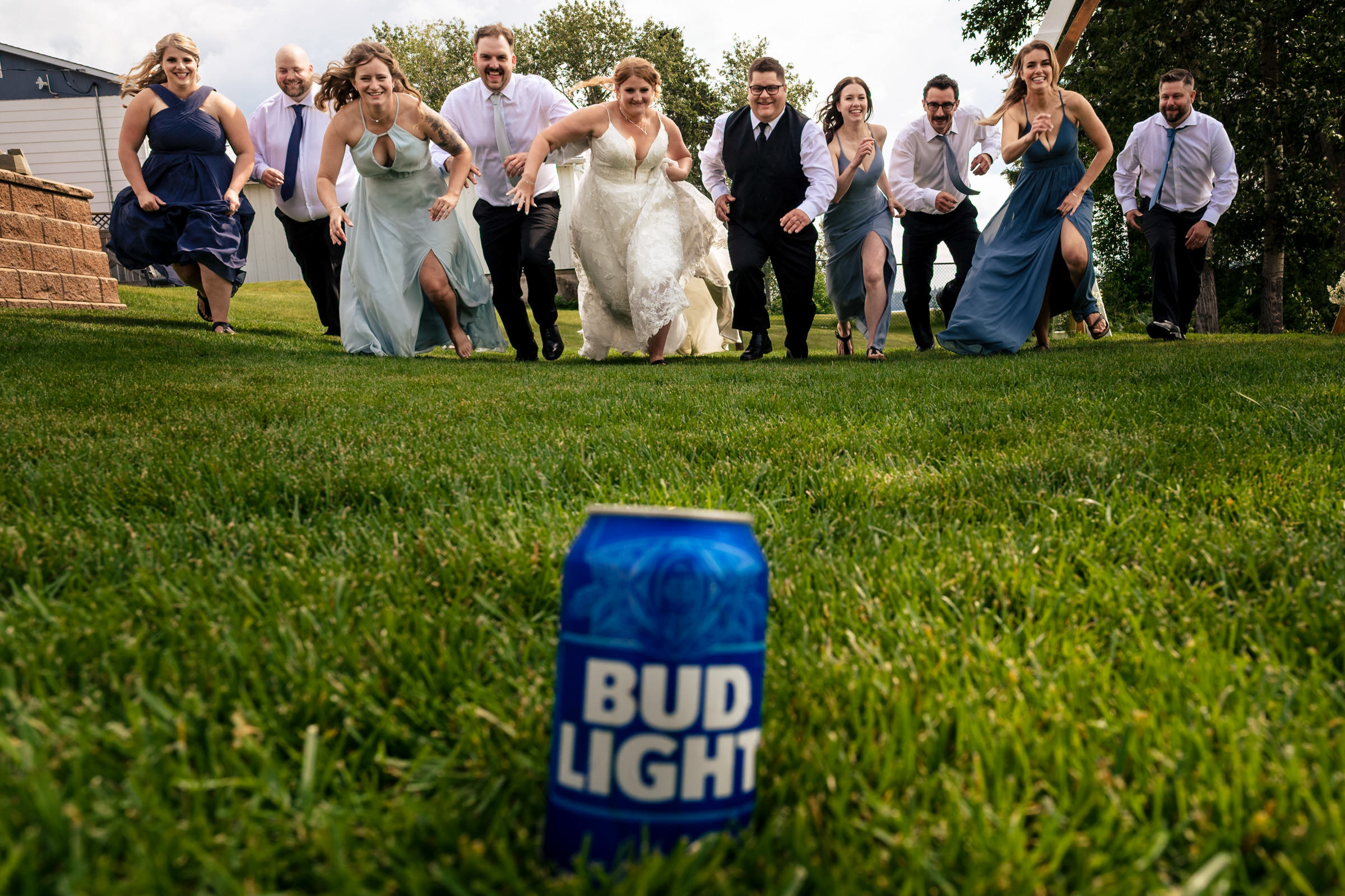 A wedding party playfully runs down a grassy hill towards the camera, capturing one of their favorite wedding photos. In the foreground, a can of Bud Light sits on the grass. The group includes bridesmaids in dresses and groomsmen in suits, with the bride and groom in the center.