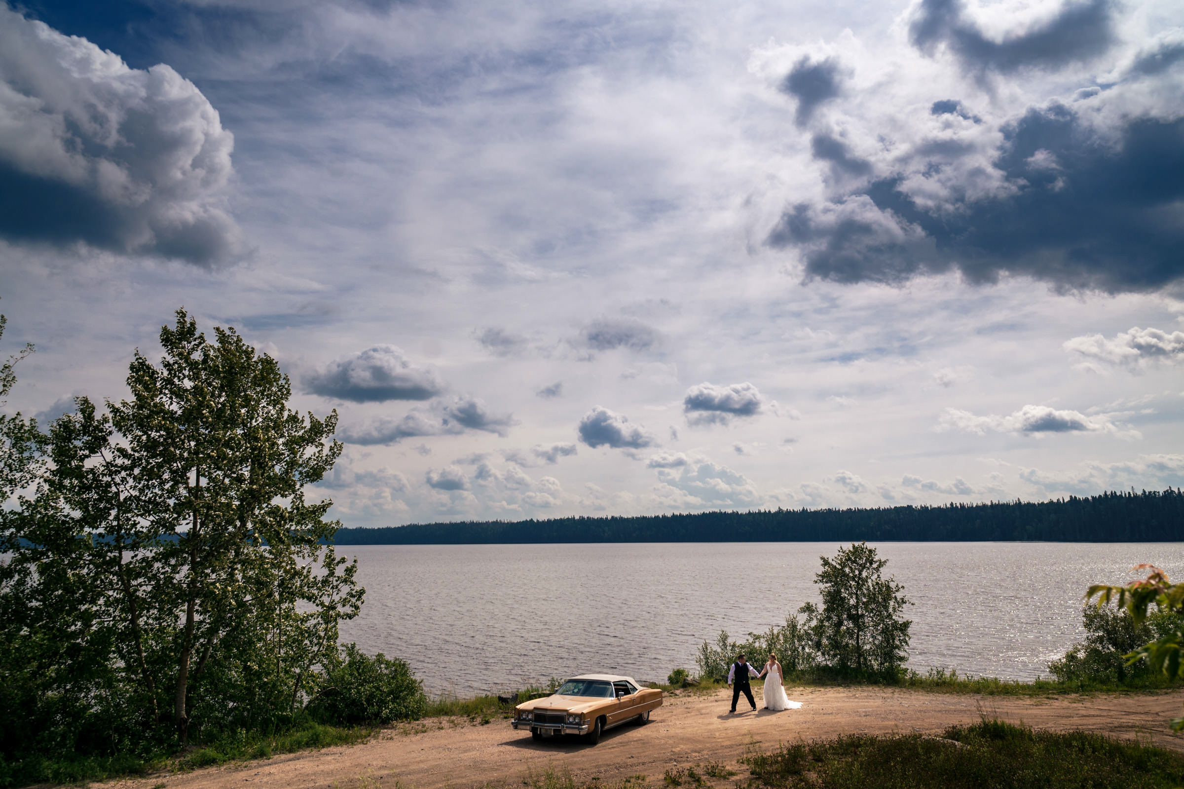 A bride and groom stand hand in hand near a vintage car on a dirt path overlooking a vast lake, creating one of their favourite wedding photos. The sky is partly cloudy, with sunlight peeking through, while trees frame the scene, adding to the romantic and picturesque atmosphere.