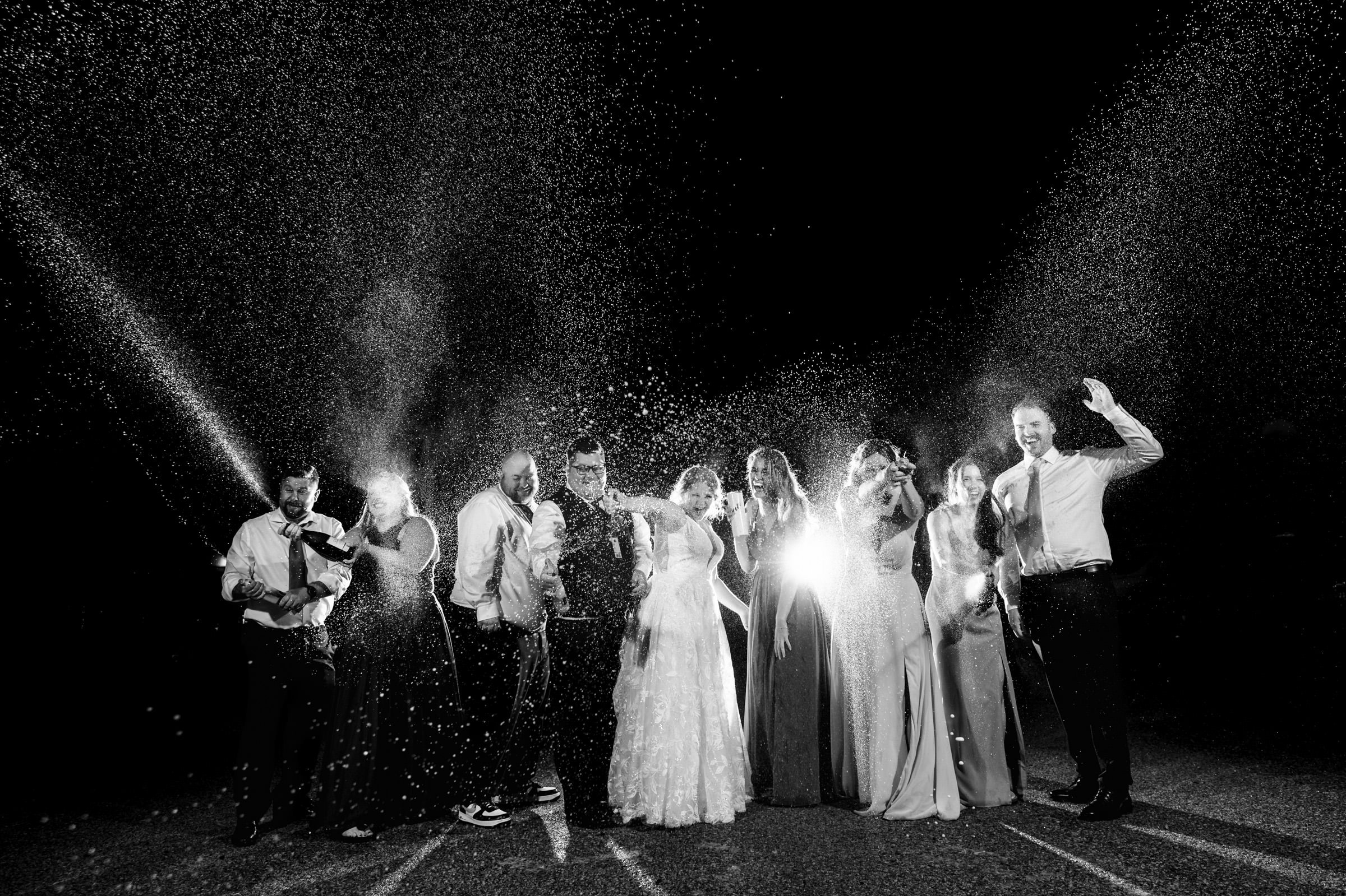 A black and white photo capturing one of the favourite wedding photos: a jubilant party celebrating at night. A group in formal attire stands in line, smiles wide as they spray champagne, creating a sparkling mist amidst the dark background.