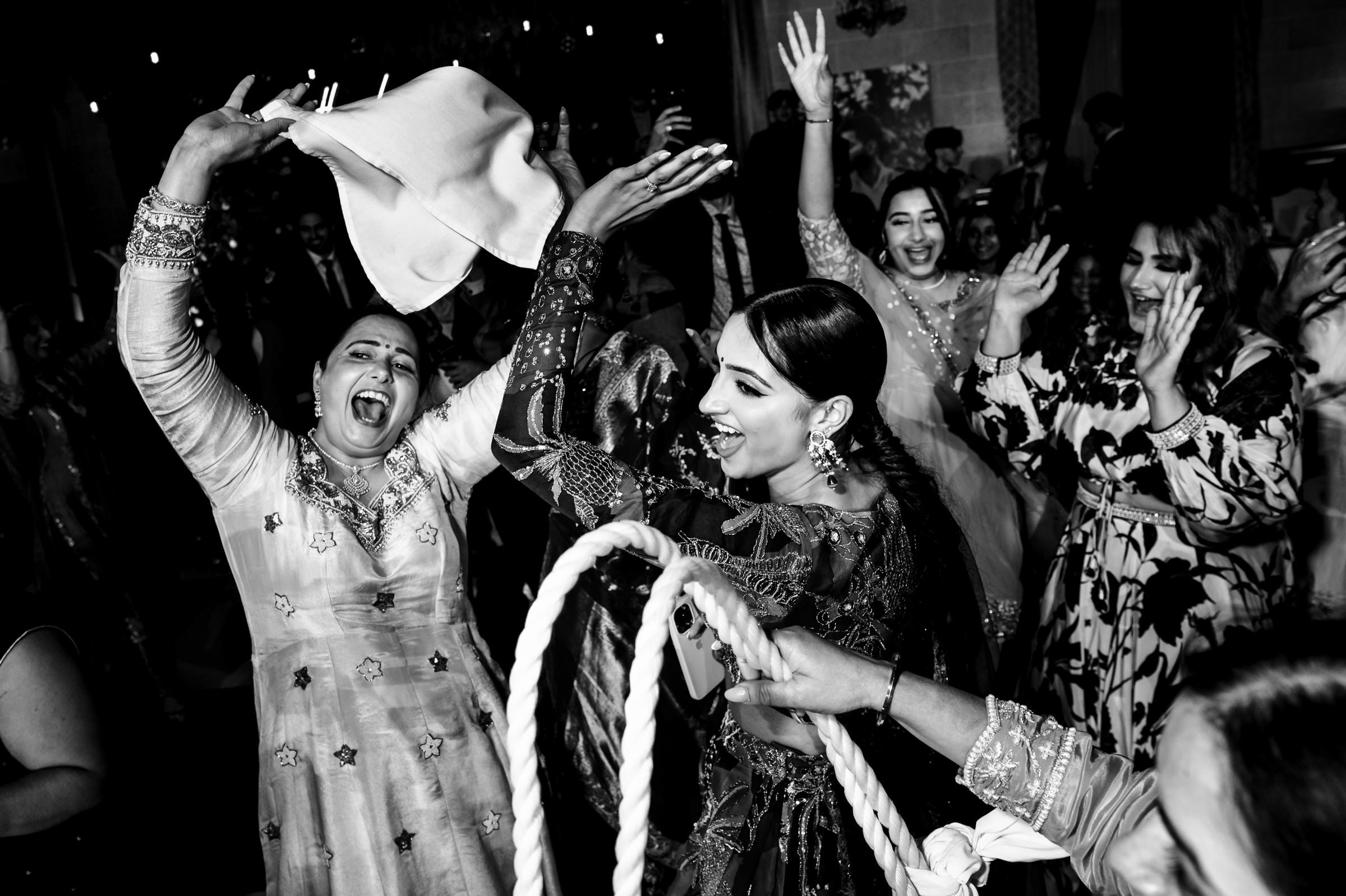 A black and white photo captures a lively group of people dancing and celebrating, reminiscent of favourite wedding photos. A woman in front joyfully holds a scarf, her exuberance matched by laughter and raised hands that echo the excitement and happiness of the festive atmosphere.