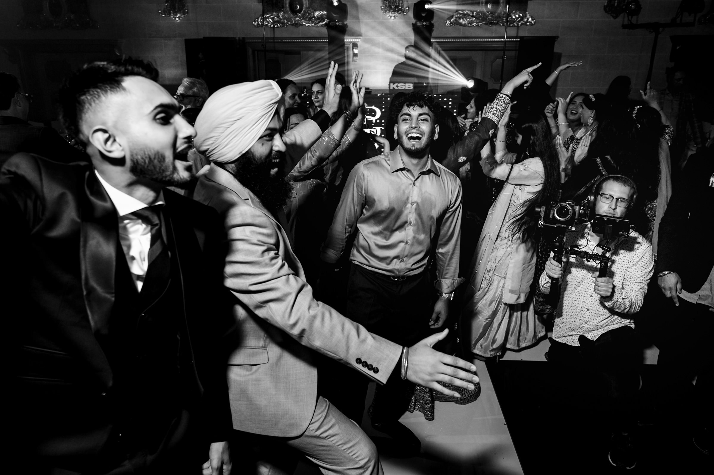 A lively group of people dancing energetically at an indoor celebration, reminiscent of favourite wedding photos. The black-and-white image captures diverse individuals in festive attire, with some smiling and others gesturing enthusiastically. A videographer films the joyous scene.