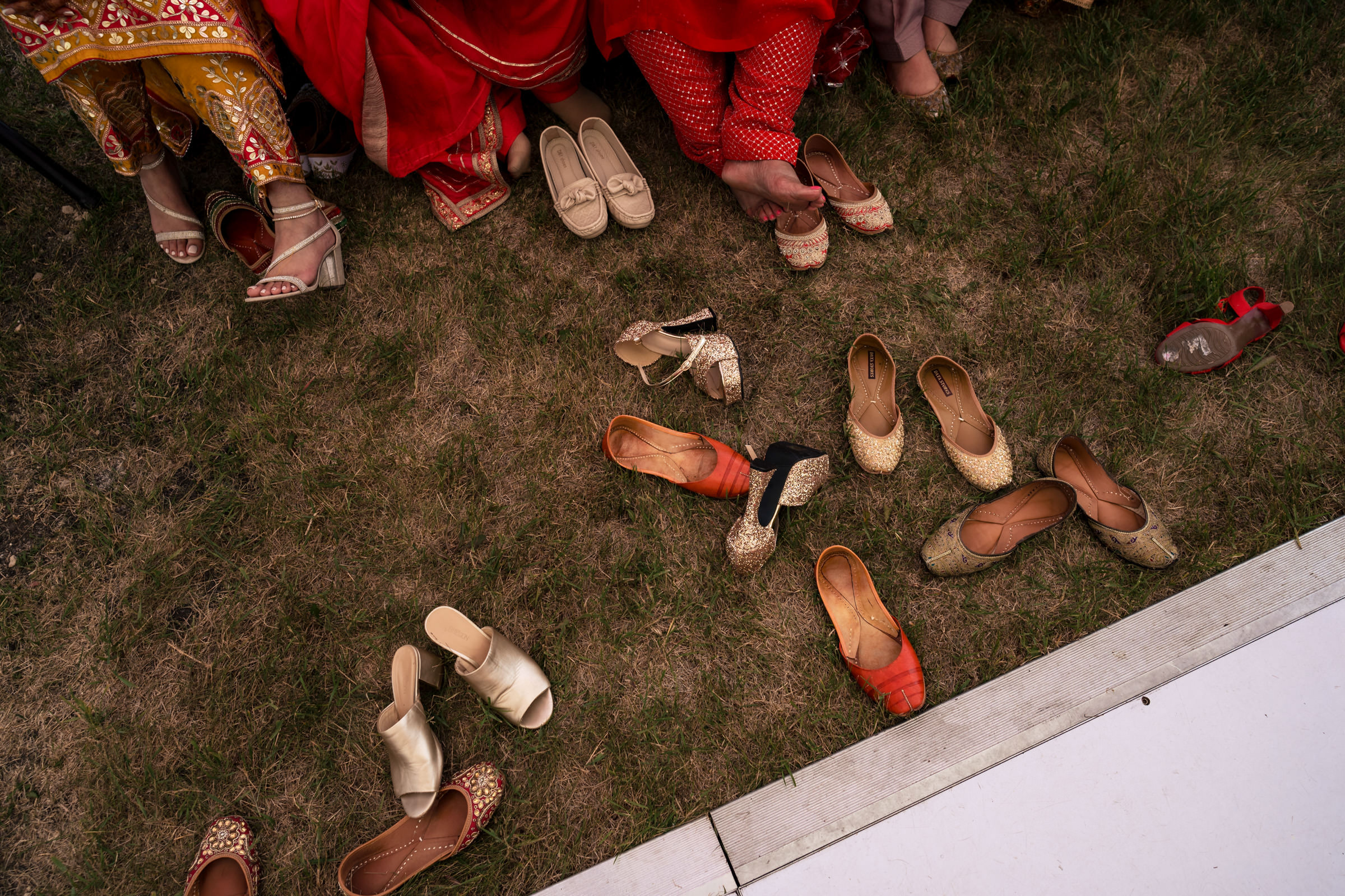 A group of people in colorful traditional attire sit on grass, surrounded by an assortment of shoes. Some reach down to adjust or select sandals, flats, or loafers in various colors, capturing moments that could easily become their favorite wedding photos.