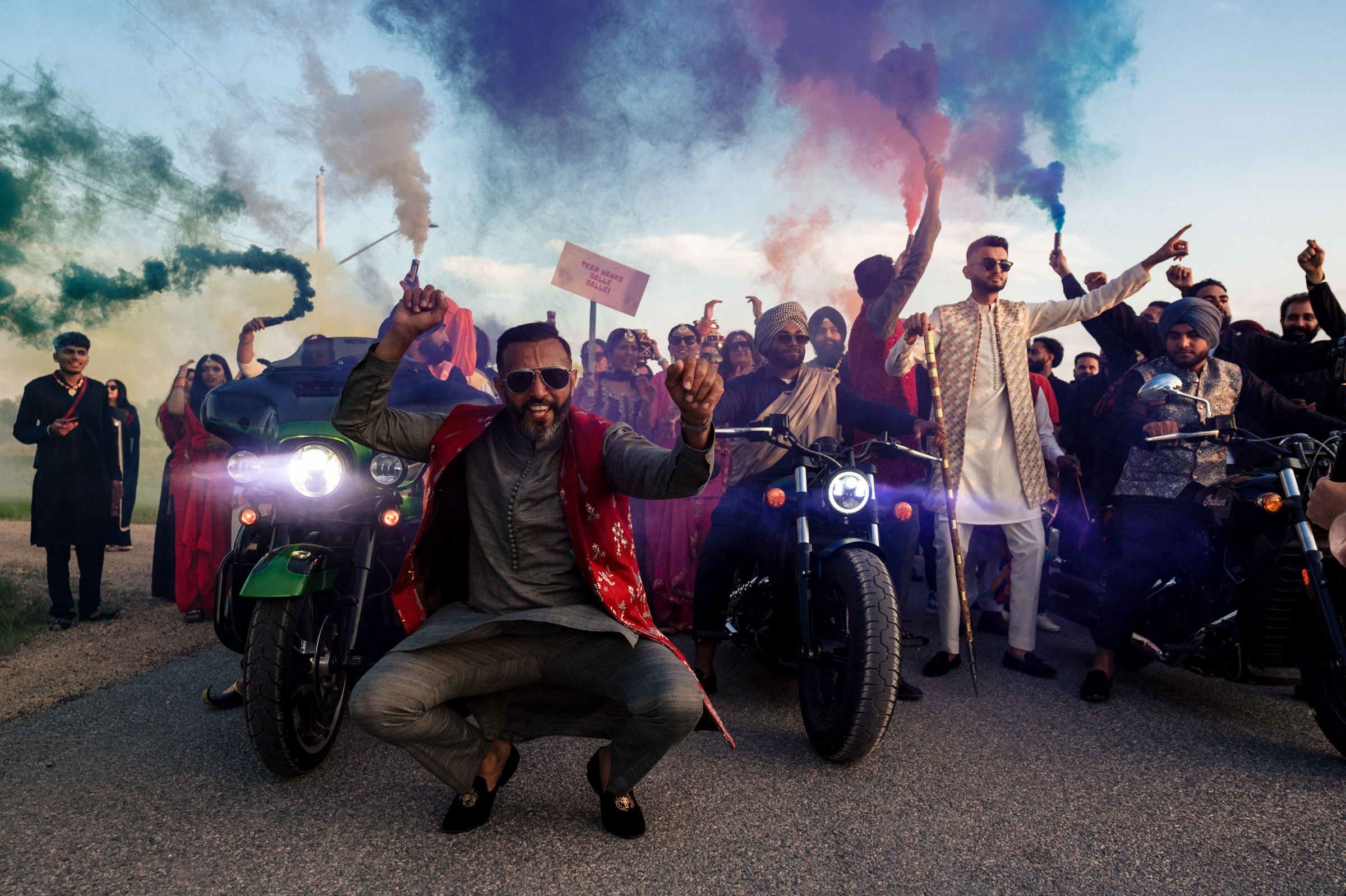 A colorful, festive scene unfolds as a group celebrates on the road, reminiscent of favorite wedding photos. Smoke flares in various colors fill the air. In the foreground, two men pose on motorcycles; one crouches and smiles at the camera, both donning vibrant attire.