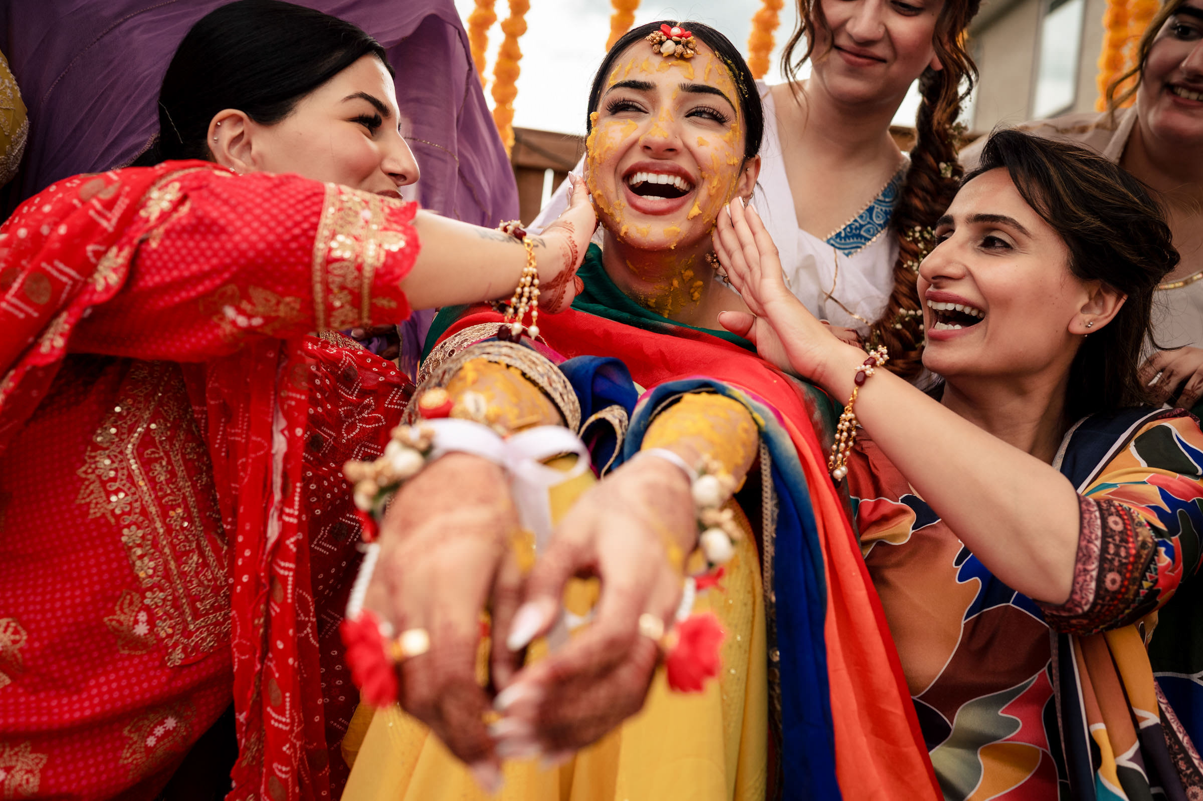 A joyful woman is seated with turmeric paste on her face and arms during a traditional ceremony, surrounded by friends wearing colorful attire. As hands lovingly apply more paste, marigold decorations add charm to the scene—one of her favorite wedding photos capturing this vibrant moment.