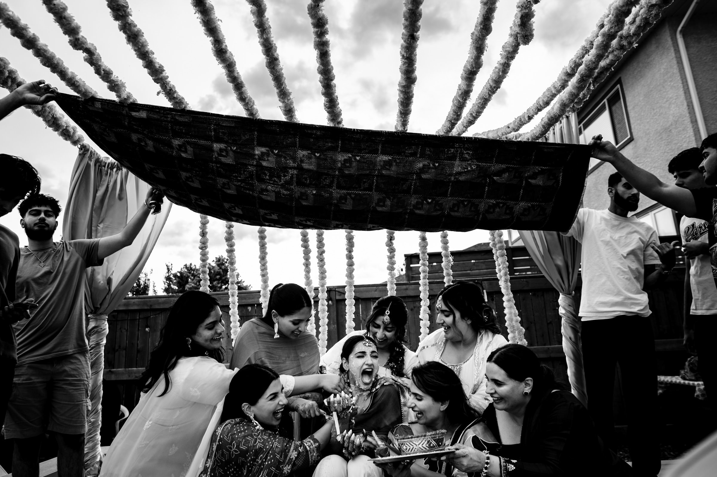 A joyful black-and-white scene captures one of the favourite wedding photos from a pre-wedding ceremony. A bride, surrounded by friends and family, laughs as they apply haldi to her face. Men hold a decorative canopy overhead against a backdrop of floral garlands and a house.