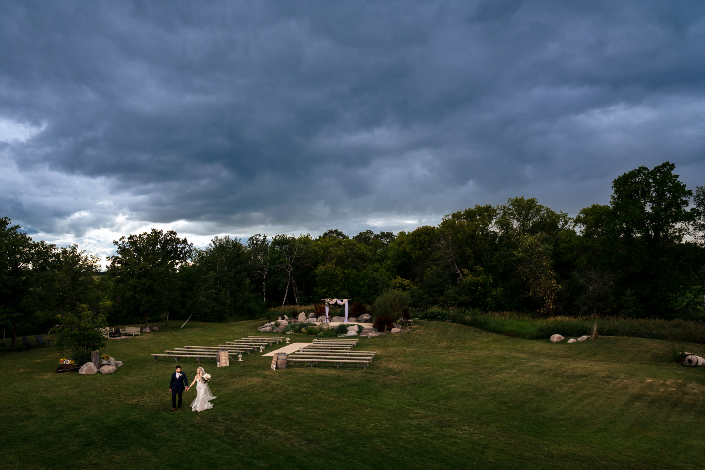 A bride and groom walk hand in hand across a grassy field under a dramatic cloudy sky, creating one of their favourite wedding photos. The outdoor setting includes rows of benches and a small archway adorned with fabric and flowers, with trees lining the background to create a serene, natural atmosphere.