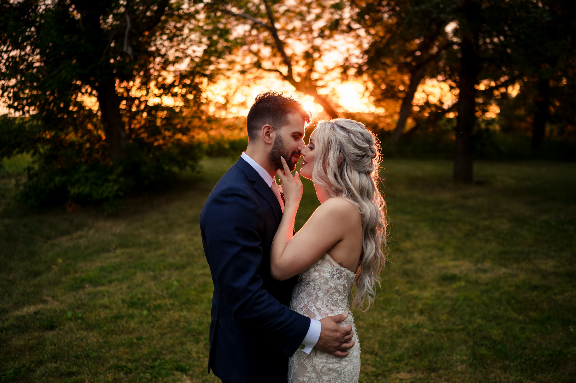 A couple embraces in a grassy field during sunset, capturing one of their favorite wedding photos. The man wears a dark suit and the woman stuns in a strapless lace gown. They gaze into each other's eyes, surrounded by trees and the warm, glowing sunlight in the background.