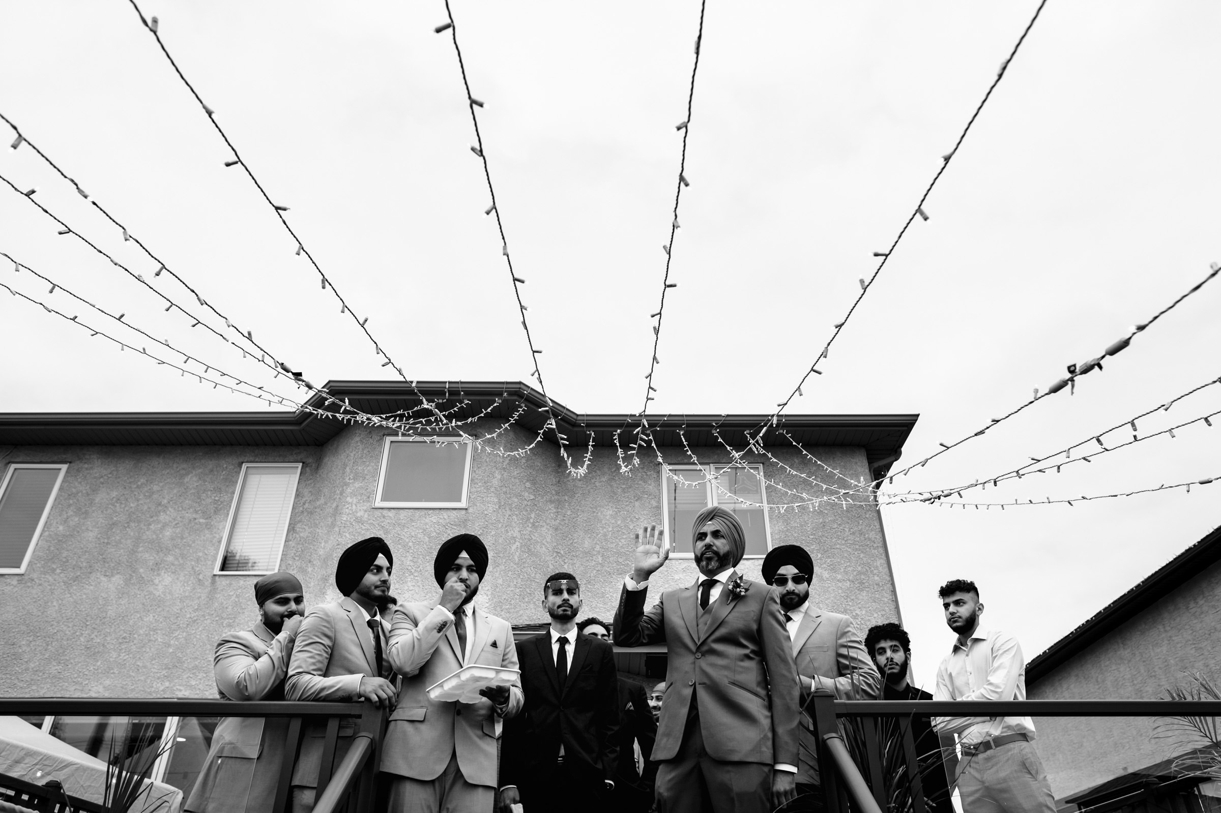 A group of men in suits stand on a deck in front of a house with string lights overhead, captured candidly in black and white. One man in a turban waves, while others listen and speak, blending seamlessly into the moment—a perfect addition to any collection of favorite wedding photos.
