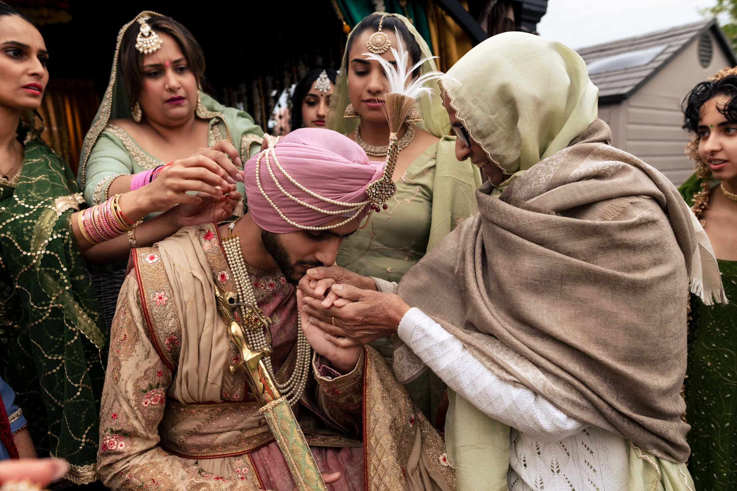 A groom in traditional attire kisses the hand of an elderly woman in a headscarf, surrounded by family members in colorful clothing. This heartfelt moment is one of my favorite wedding photos, capturing the essence of cherished family bonds and timeless traditions.