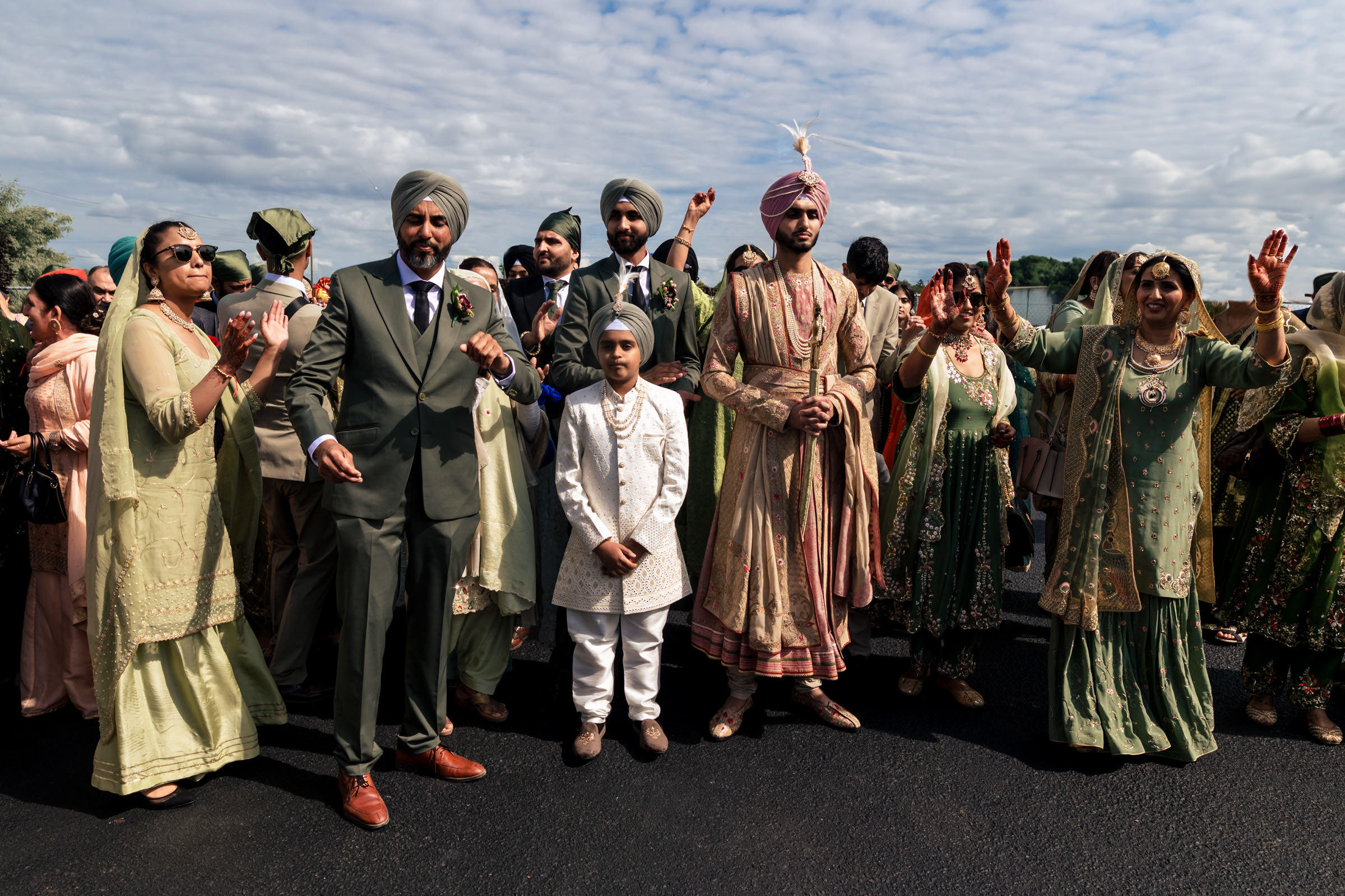 A group of people dressed in traditional Indian attire celebrate outdoors, their favourite wedding photos capturing the vibrant scene. They wear colorful and ornate clothing, including turbans, suits, and dresses. The festivities unfold joyously under a partly cloudy sky.