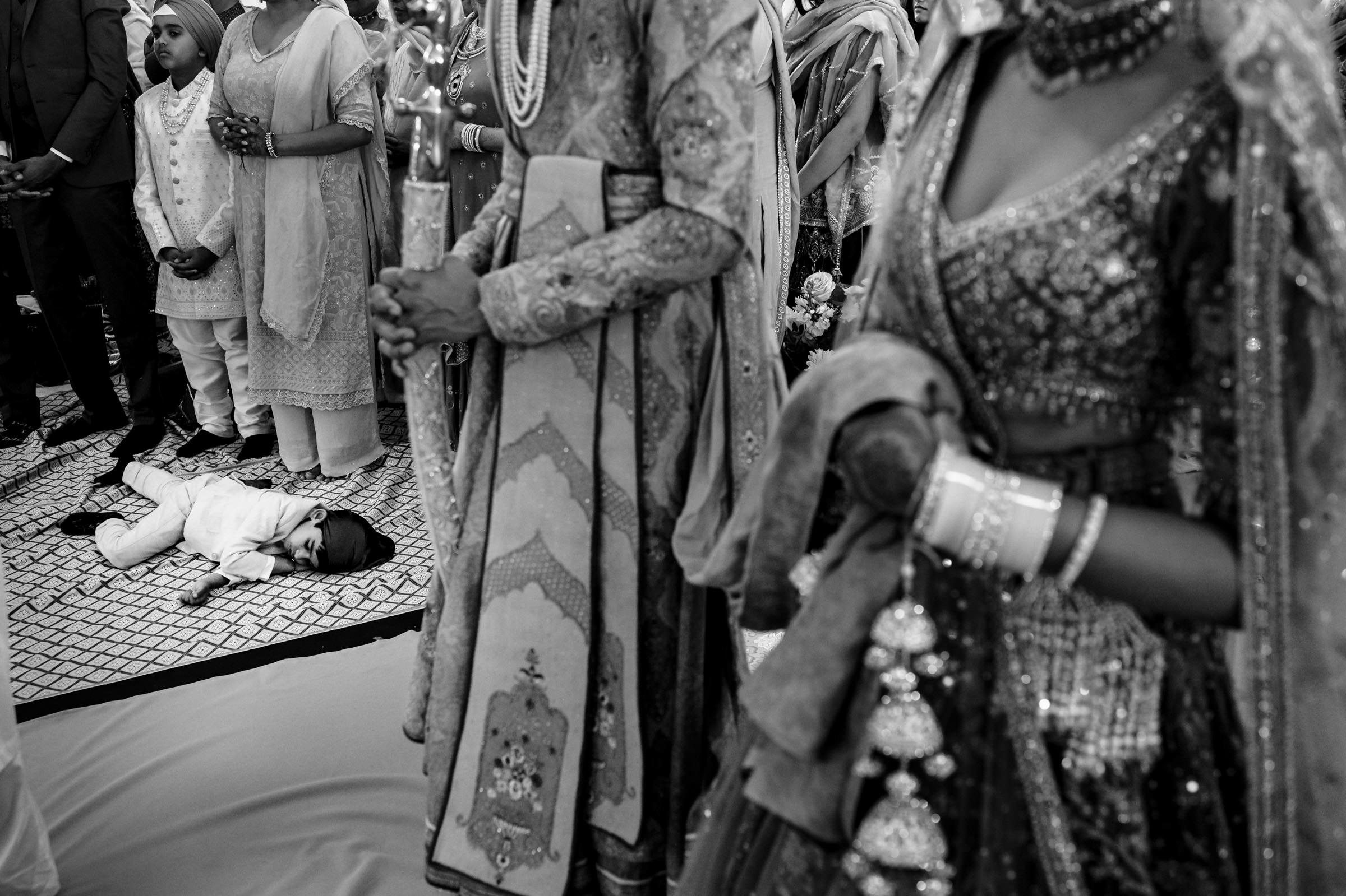 A child lies on the floor during a cultural ceremony, surrounded by adults in ornate traditional clothing, capturing one of my favourite wedding photos. The scene is busy, with intricate patterns and textures visible on the garments, creating a lively and ceremonial atmosphere.