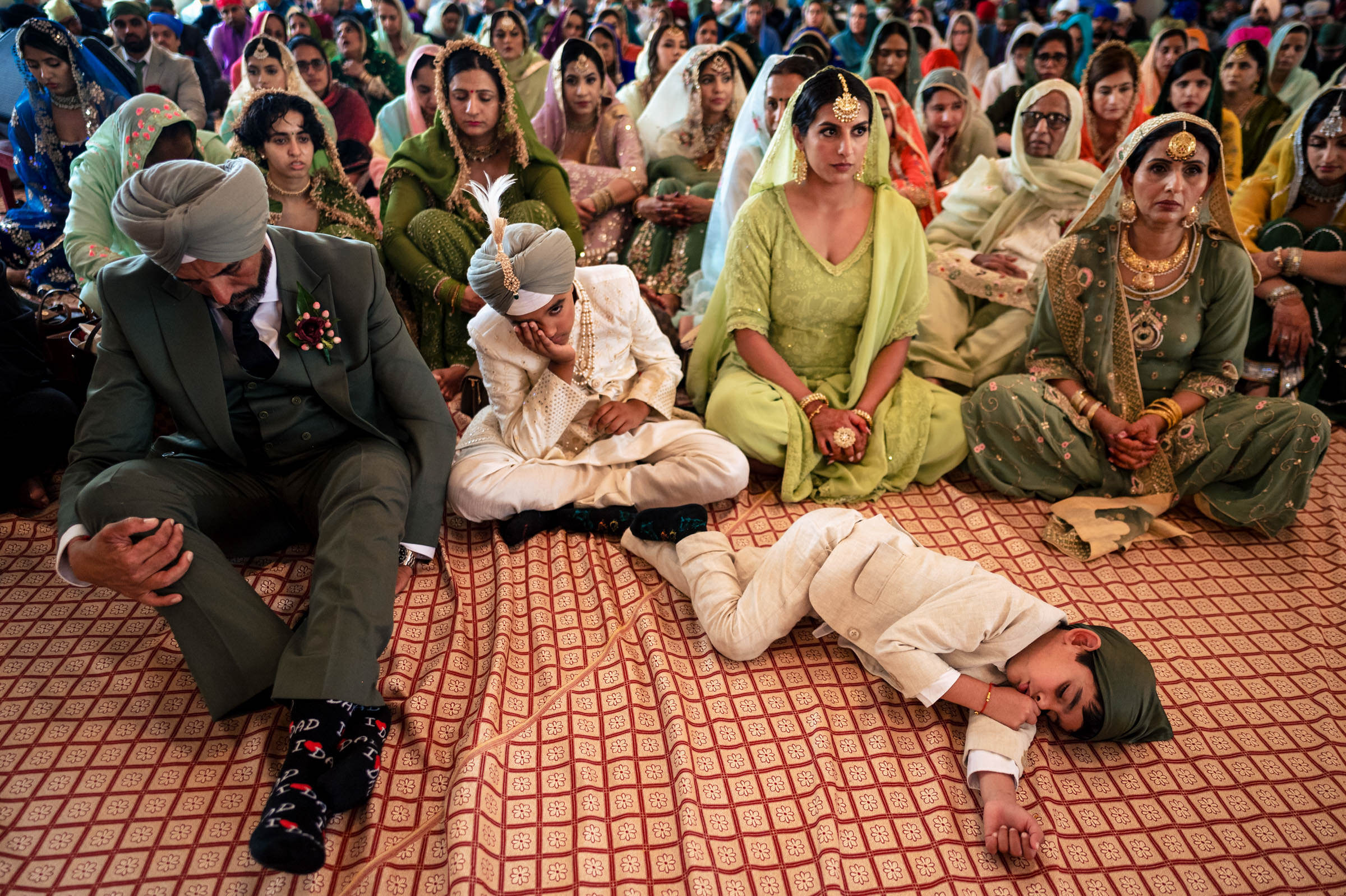 A family dressed in traditional attire sits on a patterned floor, capturing one of their favourite wedding photos. A child in a light-colored outfit lies down while another sits cross-legged, looking down. The adults around them are seated, observing the event with various expressions.