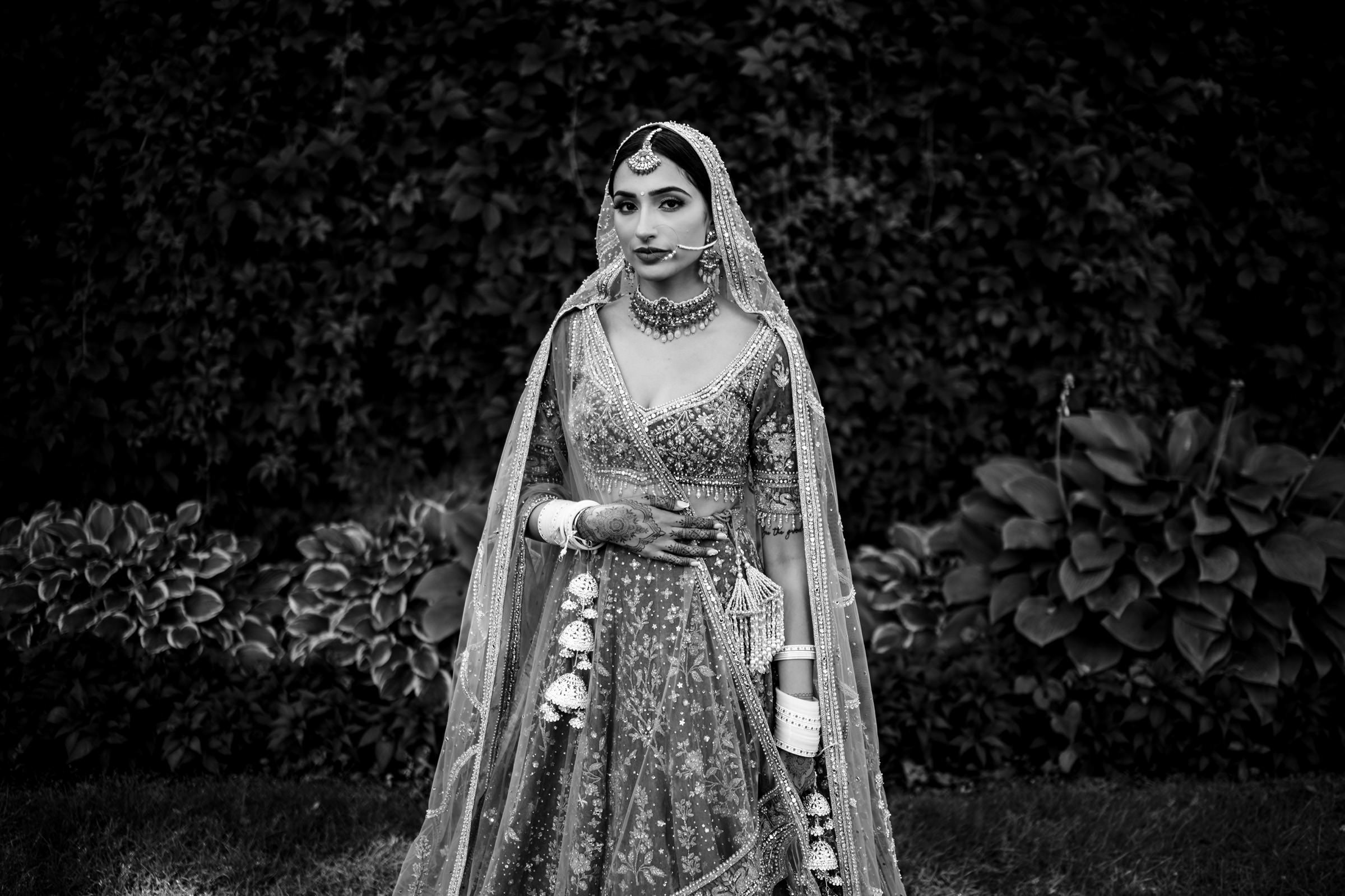 A bride in an ornate traditional outfit stands outdoors, showcasing her detailed jewelry, including a headpiece, necklace, and bangles. Captured in black and white as one of her favorite wedding photos, she holds her garment with henna-decorated hands against a backdrop of lush foliage.