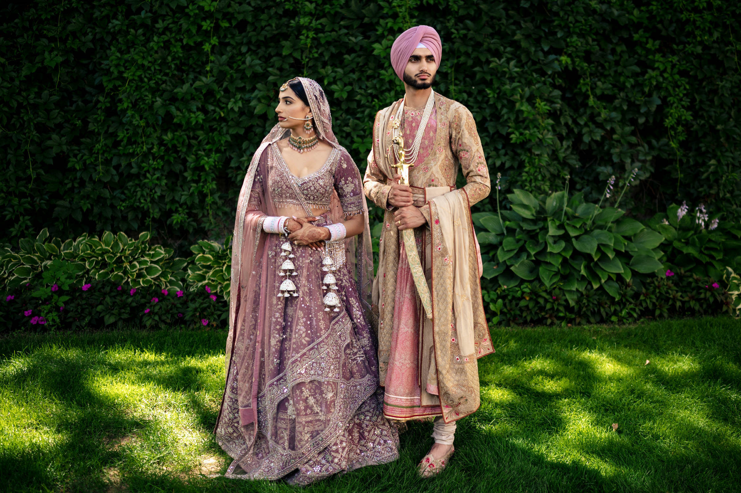 A couple poses thoughtfully outdoors in their favourite wedding photo, surrounded by lush greenery. The woman dazzles in an ornate pink lehenga, while her partner complements her in a matching sherwani, holding a sword. Together, they embody the grace of traditional South Asian attire.