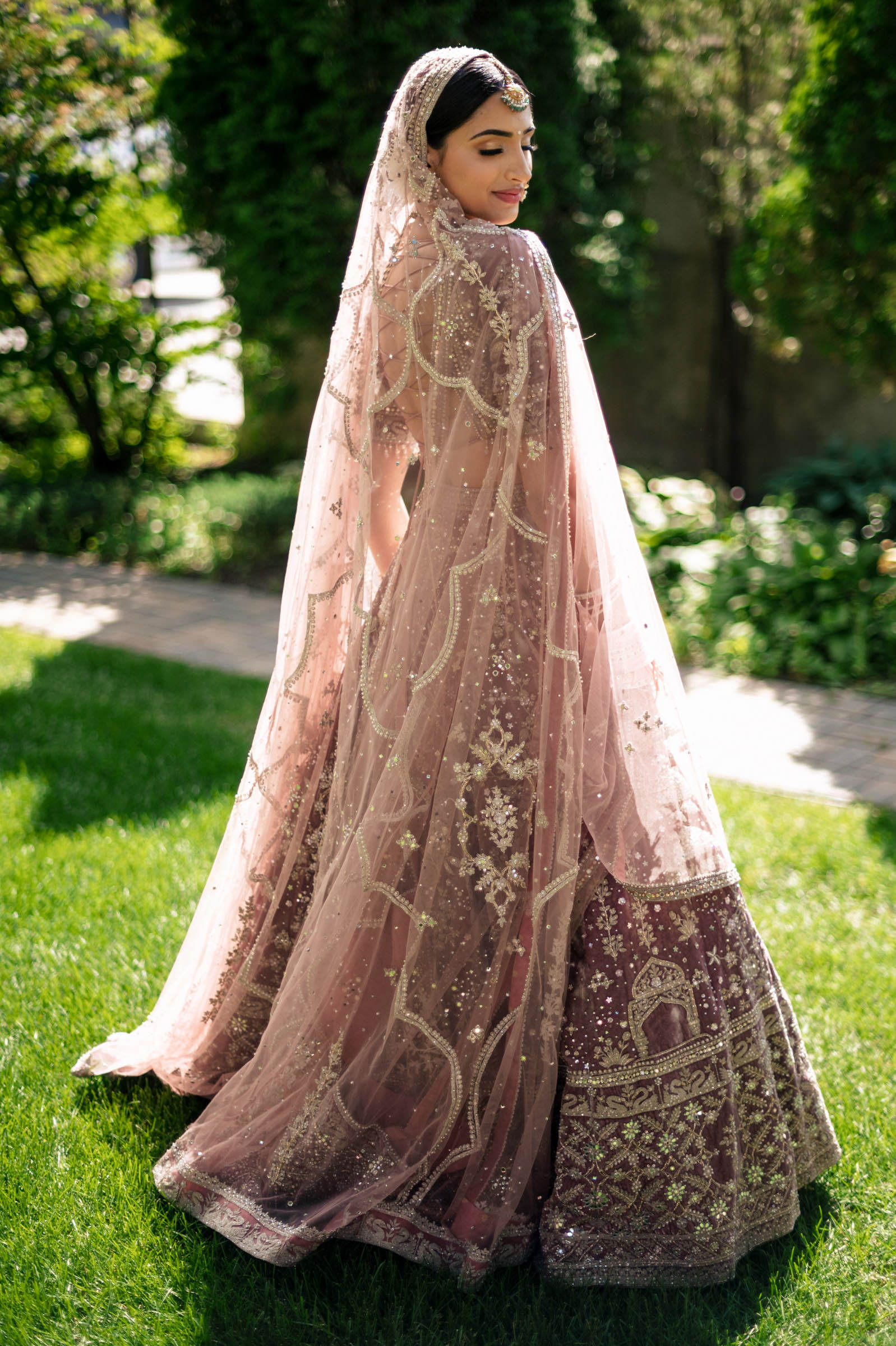 A woman in embroidered, sheer veil and traditional attire stands on a grassy path surrounded by greenery, looking over her shoulder. Her dress features intricate patterns and beadwork. Sunlight filters through the trees, creating a soft, warm ambiance—capturing one of my favorite wedding photos.