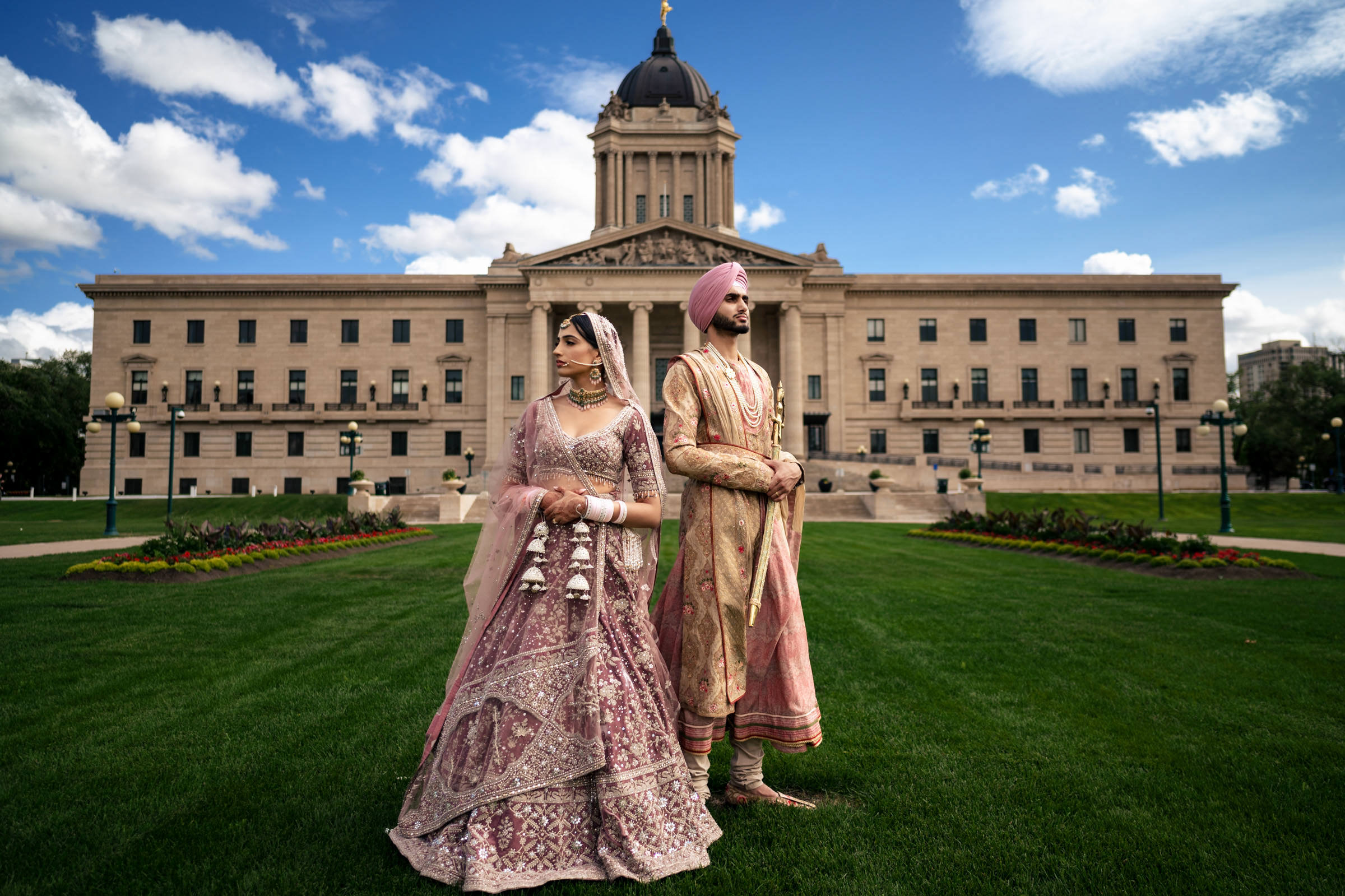A couple in traditional South Asian wedding attire stands on a lawn before a historic building, capturing one of their favorite wedding photos. The woman dazzles in an ornate pink and silver lehenga, while the man sports a matching sherwani and turban against a partly cloudy sky.