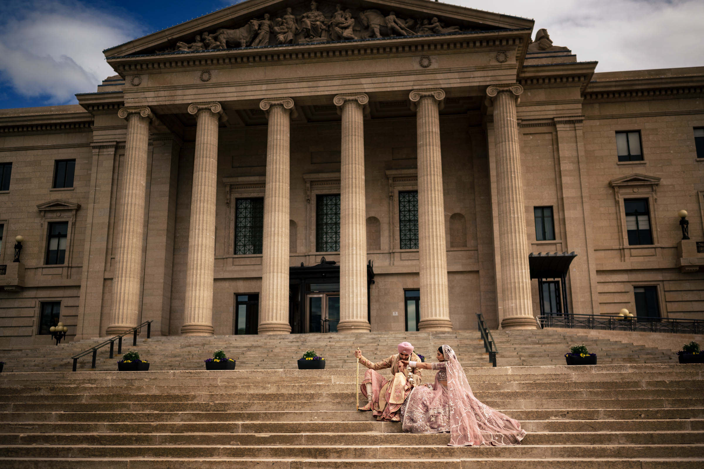 A couple in traditional attire sits on the steps of a grand building with tall columns, capturing one of their favourite wedding photos. The groom wears a turban, and the bride an ornate gown with a veil. The partly cloudy sky provides a dramatic backdrop for the historic architecture.